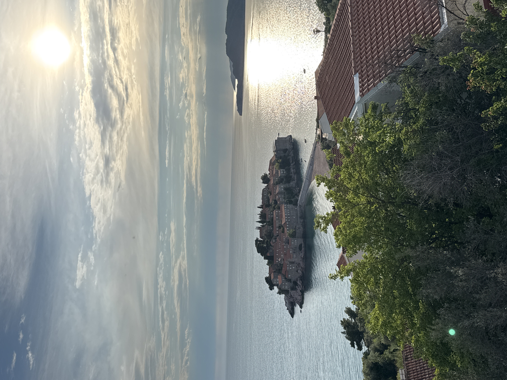 The image presents a vertically oriented view of Sveti Stefan, Montenegro, at what appears to be dusk. The sky, filled with striated clouds, holds a bright sun at its upper left. The sea reflects the sun's glare, creating a shimmering effect. In the center of the image, an island with red-roofed buildings is connected to the mainland by a narrow causeway. This island, known as Sveti Stefan, features dense clusters of structures. The right side of the image contains buildings with red tile roofs partially obscured by trees with green foliage. The environment suggests a coastal, potentially elevated, vantage point.