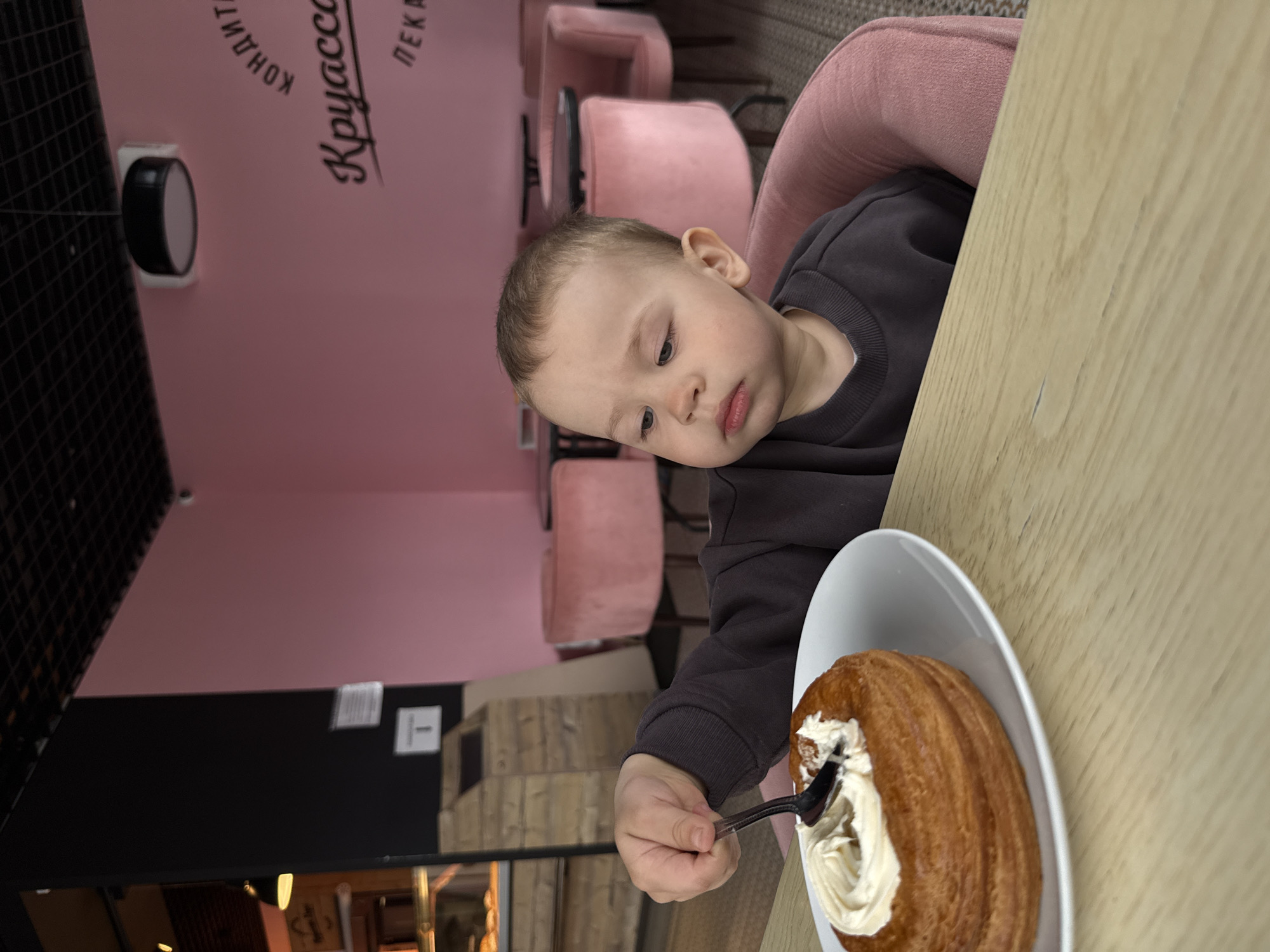 The image, rotated counter-clockwise, depicts a young child sitting at a table in a modern cafe or bakery. The child, likely a toddler or preschooler with light hair, is wearing a dark grey long-sleeved top and is seated in a plush, light pink upholstered chair. They are looking down with a focused expression, holding a small black spoon, about to eat a pastry.
On a white plate before the child is a round, golden-brown baked good with a ribbed texture, possibly a cronut or similar pastry, topped with a generous swirl of white cream or frosting. The table surface is light wood.
The background features a vibrant pink wall with dark Cyrillic text, which includes partial words like "КОНДИТ" (likely "Кондитерская" - Confectionery), "Круасса" (likely "Круассан" - Croissant), and "ПЕКА" (likely "Пекарня" - Bakery), indicating the type of establishment. Below the pink wall, a dark panel is visible, and further into the background, more pink chairs and a dark, grid-like structure suggest cafe seating and decor. The scene captures a peaceful moment of a child enjoying a treat in a well-lit, contemporary interior in Rusanovo, Russia.