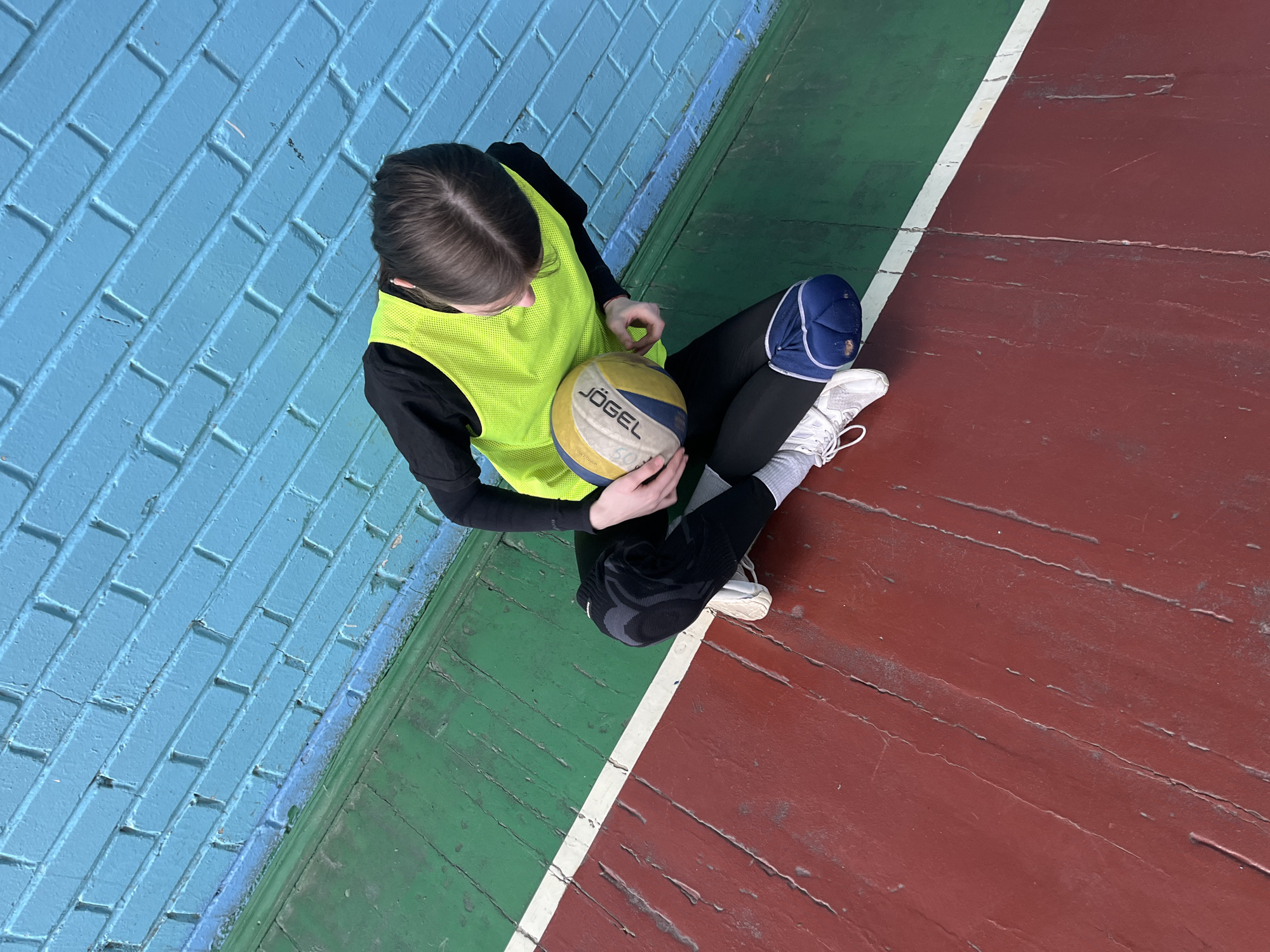 An overhead shot captures a person sitting on a sports court, likely in an indoor gym. The individual is dressed in athletic wear, including a black long-sleeved shirt, a bright neon yellow sports vest, black leggings, white athletic shoes, and white socks. They have knee pads on both legs; one is blue and white, while the other is black with a grey pattern.
The person is holding a volleyball that is yellow, white, and blue, with the brand name "JÖGEL" prominently displayed. Their posture suggests a moment of rest or contemplation.
The court floor is distinctly painted in two primary colors: a dark red section and a dark green section, separated by a worn white line. The floor shows signs of heavy use with visible cracks and scuff marks. In the background, a brick wall is painted a vibrant light blue, further indicating an indoor setting. The overall environment appears to be an older, well-used sports facility. This scene is located in Saint Petersburg, Russia.