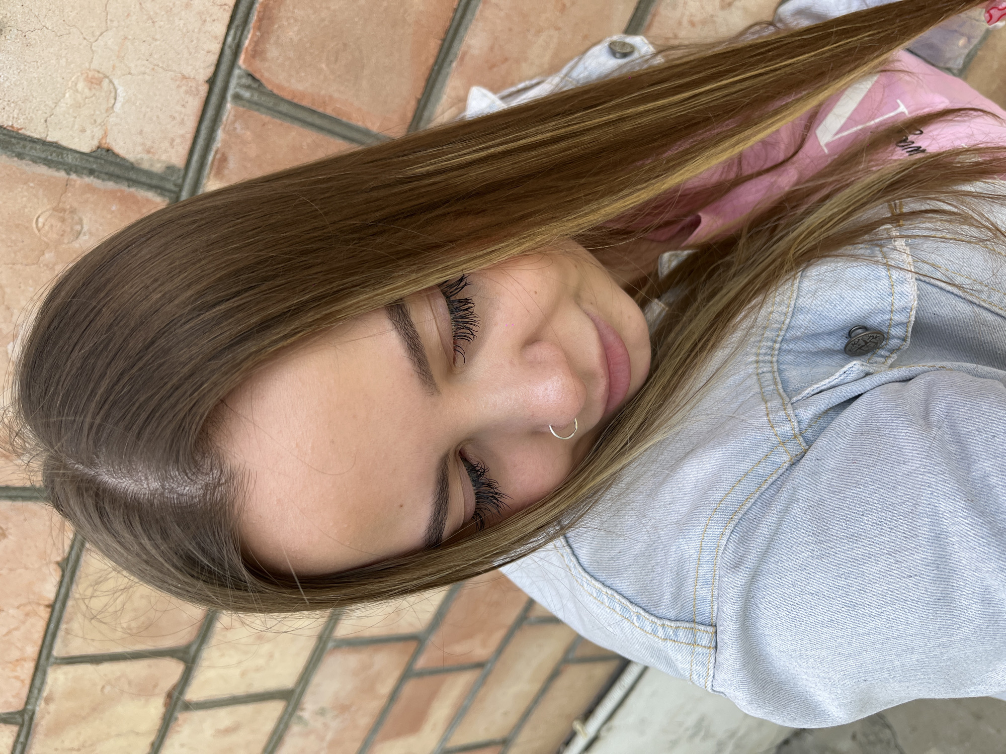 A young woman poses for a photo with a brick wall in the background. She is wearing a light blue denim jacket and a pink shirt. The image was taken outdoors. The location is the city of Novopokrovskaya, Russia.