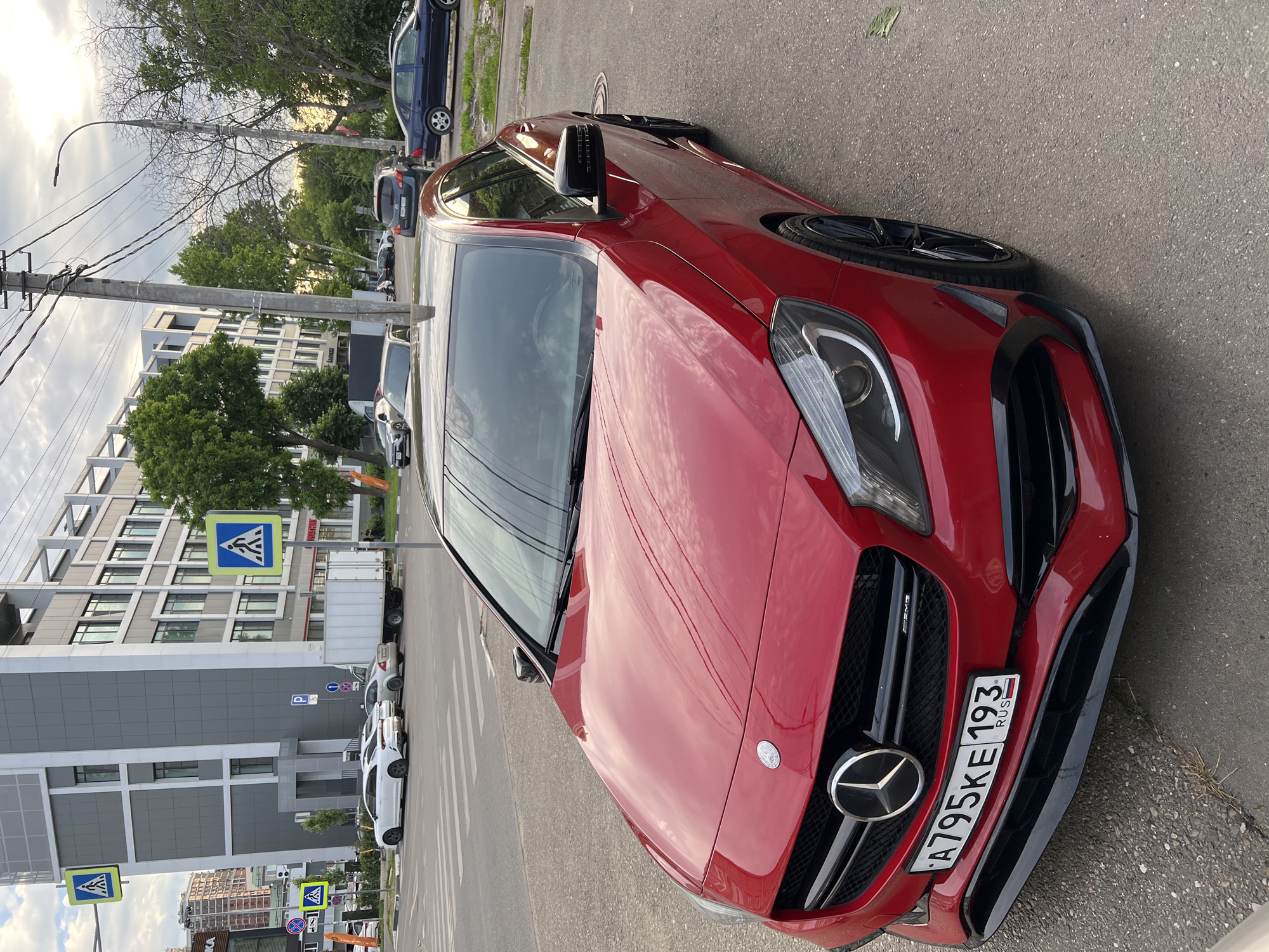 A red Mercedes-Benz car, possibly an AMG model indicated by a small badge on the grille, is parked on the side of a street. The car is oriented with its front facing the bottom right of the image, showing its sleek design, prominent Mercedes-Benz emblem on the grille, and multi-spoke black wheels. The license plate, visible on the front bumper, reads "A795KE 193 RUS".
The scene depicts an urban environment during what appears to be daytime, under an overcast sky. The road surface is asphalt, and there's a sidewalk or curb next to the parked car. In the background, modern multi-story buildings with a gray and white facade are visible, suggesting a city or developed area. Numerous trees with green foliage line the street, some appearing dense while others are sparser, indicating late spring or summer.
Utility poles with power lines stretch across the frame, a common feature in urban landscapes. Other vehicles, including sedans, SUVs, and a white truck, are parked along the street further down or are partially visible. Several blue and white pedestrian crossing signs are noticeable, along with other street signs including a "P" for parking. The overall impression is a typical street scene in a city, with the red Mercedes-Benz being the central focus. No people are explicitly visible in the frame.