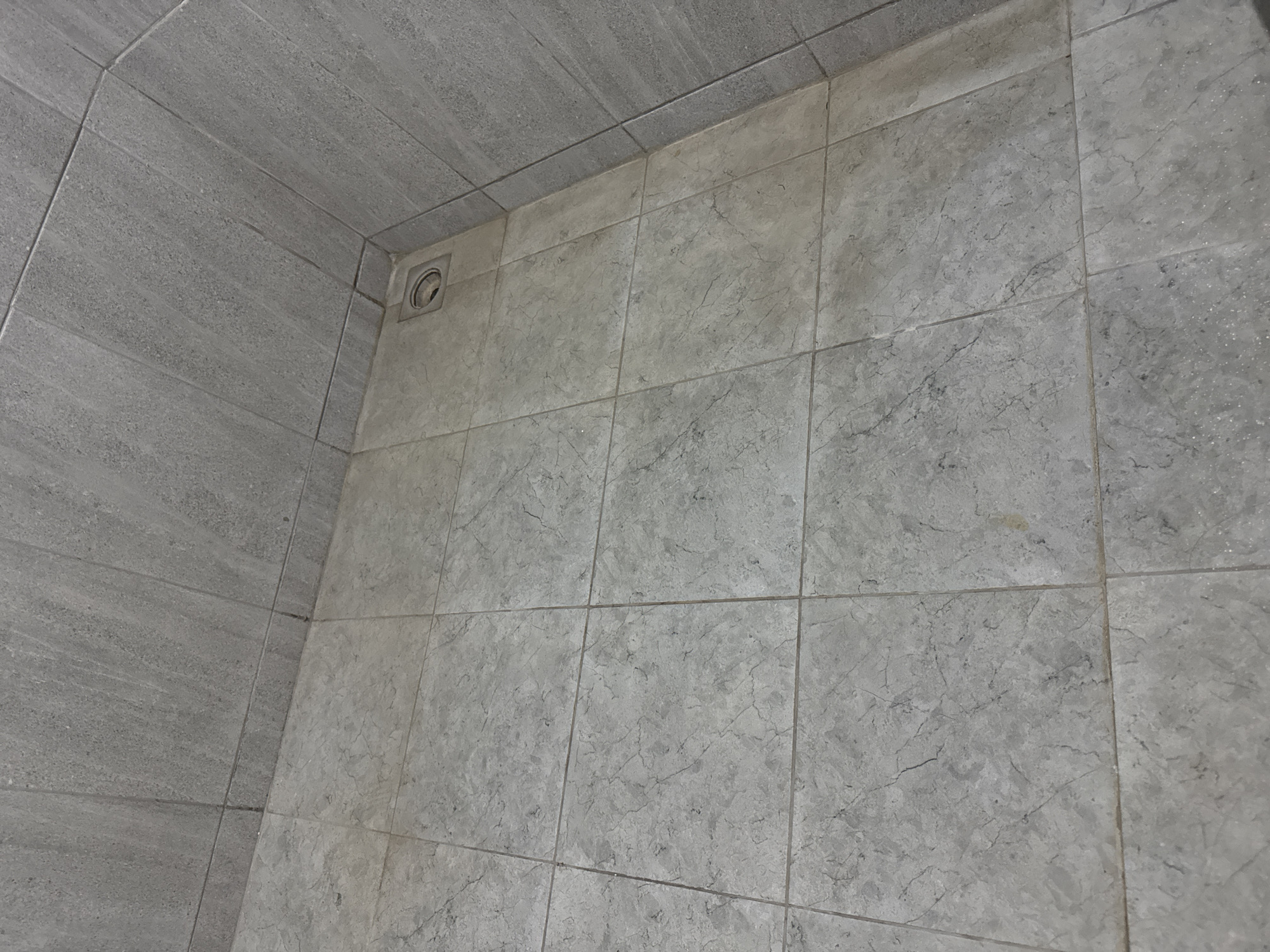 The image shows a close-up view of a tiled corner, likely in a bathroom or shower area. The tiles are a light gray color with a subtle marble-like pattern. The floor tiles are square, and the wall tiles are rectangular and oriented vertically. In the corner where the floor and walls meet, there is a square drain with a circular grate. The lighting suggests an indoor environment. There is no indication of the city of Sukawati, Indonesia, in the image itself.