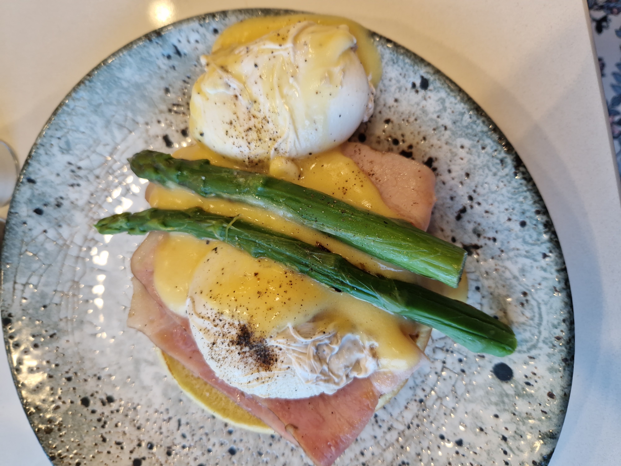 A close-up, top-down shot shows a plate of eggs Benedict with ham and asparagus. The dish is presented on a speckled grey and blue ceramic plate. Two poached eggs, generously coated in hollandaise sauce, sit atop slices of ham and what appears to be an English muffin base. Three stalks of bright green asparagus are arranged vertically in the center of the dish, also covered in sauce. Black pepper is sprinkled over the eggs. The lighting is bright, and the background is out of focus, suggesting an indoor setting. There are no people or identifiable landmarks visible in the image. The dish appears to be a breakfast or brunch item.
