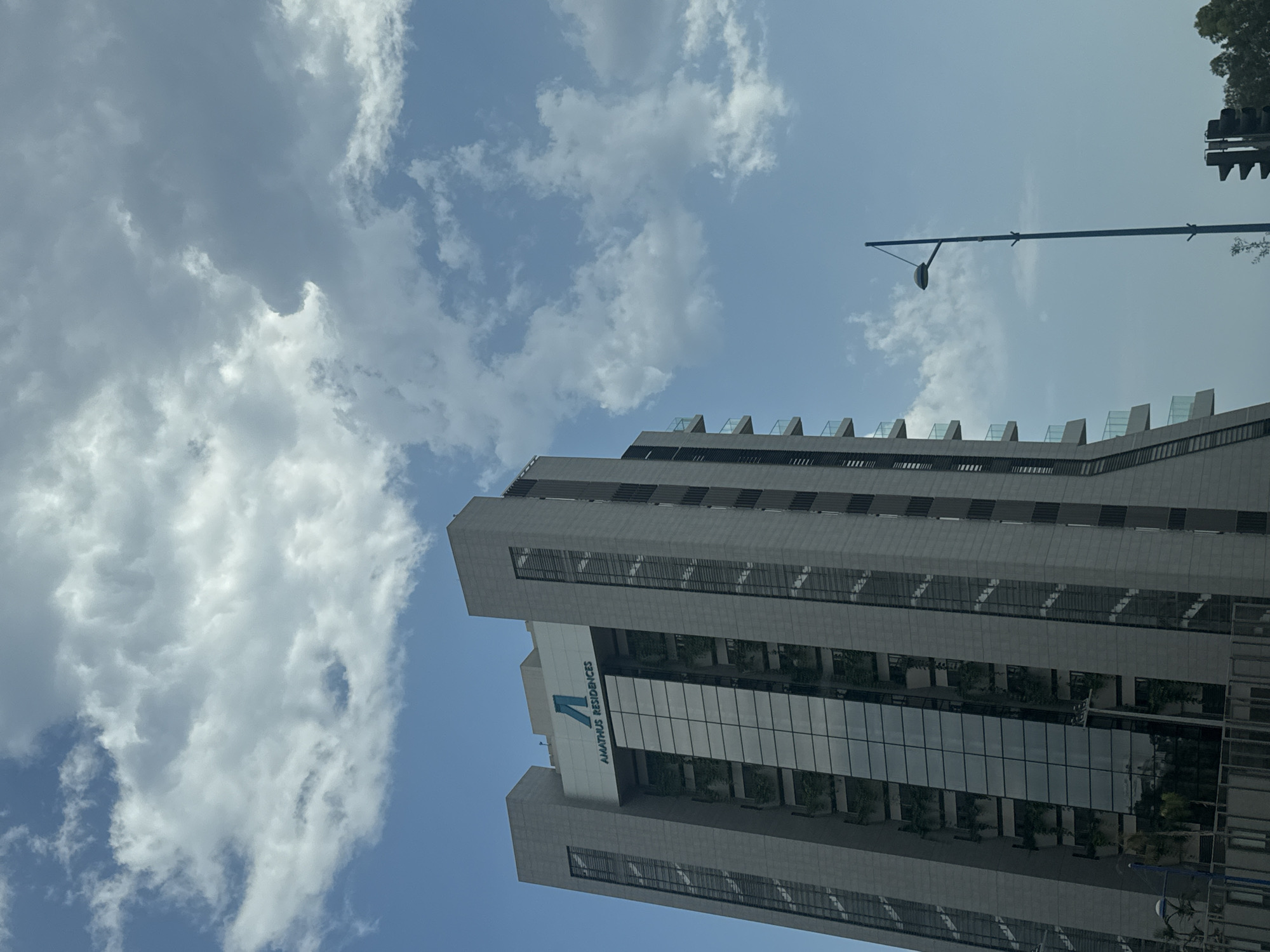 The image shows the Amathus Residences building, a modern high-rise located in the Agios Tychona community, Cyprus. The building is characterized by its gray facade, large glass windows, and balconies with glass railings. The Amathus Residences logo, featuring a blue stylized "A", is visible at the top of the building.
The sky above is a clear blue, dotted with fluffy white clouds. In the foreground, a tall blue lamppost stands to the right of the building, and a traffic light is partially visible at the bottom right corner. The surrounding environment appears to be an urban setting with trees in the distance. The overall scene captures a bright, sunny day in a coastal city.