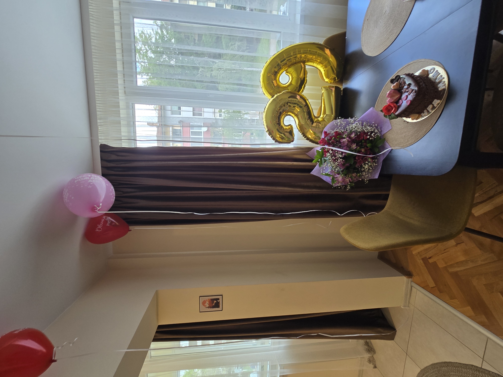 The image displays an indoor scene, likely a dining area, set up for a birthday celebration. No people are visible.
The primary subjects include festive decorations: large gold foil balloons clearly forming the number "26," a chocolate cake, and a bouquet of fresh flowers. The cake is round and chocolate-covered, placed on a golden circular stand on a dark dining table. It's decorated with a red rose and what appear to be heart-shaped chocolate pieces or strawberries. Alongside the cake is a bouquet of red and pink flowers, wrapped in light purple paper.
Two chairs, one mustard-yellow/olive green, are pulled up to the table. In the background, a large window reveals a bright daytime setting with trees and residential buildings outside, suggesting an urban or suburban location. The window is adorned with sheer white curtains and heavier brown drapes. Near the ceiling, several latex balloons are floating, including a pink one and a red heart-shaped balloon. The flooring is a wooden parquet pattern. A small framed picture is visible on a light-colored wall below the window.
The visible text includes the number "26" on the gold balloons. On the pink latex balloon, partial text "С ДНЕМ РОЖДЕ..." (likely "Happy Birthday" in Russian) is legible. The red heart-shaped balloon displays "Обичам те" (I love you in Bulgarian). Partial text "ДНЕ... НАЙ..." can be seen on the golden base of the cake. The scene clearly depicts a celebratory event, likely a 26th birthday, characterized by thoughtful arrangements and decorations, and conveyed in a bright, daytime setting.