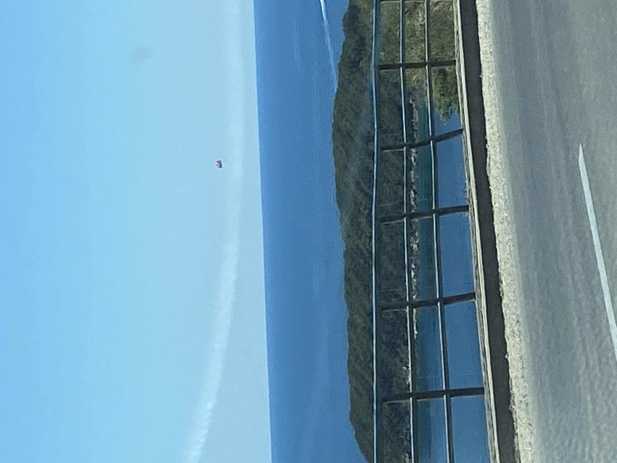 In the city of Prijevor, Montenegro, a parasailer is seen in the sky above the Adriatic Sea. The parasailer, a small red object, is suspended in the air against a clear blue sky. Below the parasailer, the deep blue sea stretches to the horizon. In the middle ground, a lush green island covered in trees is visible, bordered by the sea. A black metal railing runs across the foreground, partially obscuring the view of the island and the water. A paved road, possibly part of a street or highway, is visible in the lower portion of the image, with a white line marking a lane. The scene appears to be a scenic overlook or a point along a coastal road.