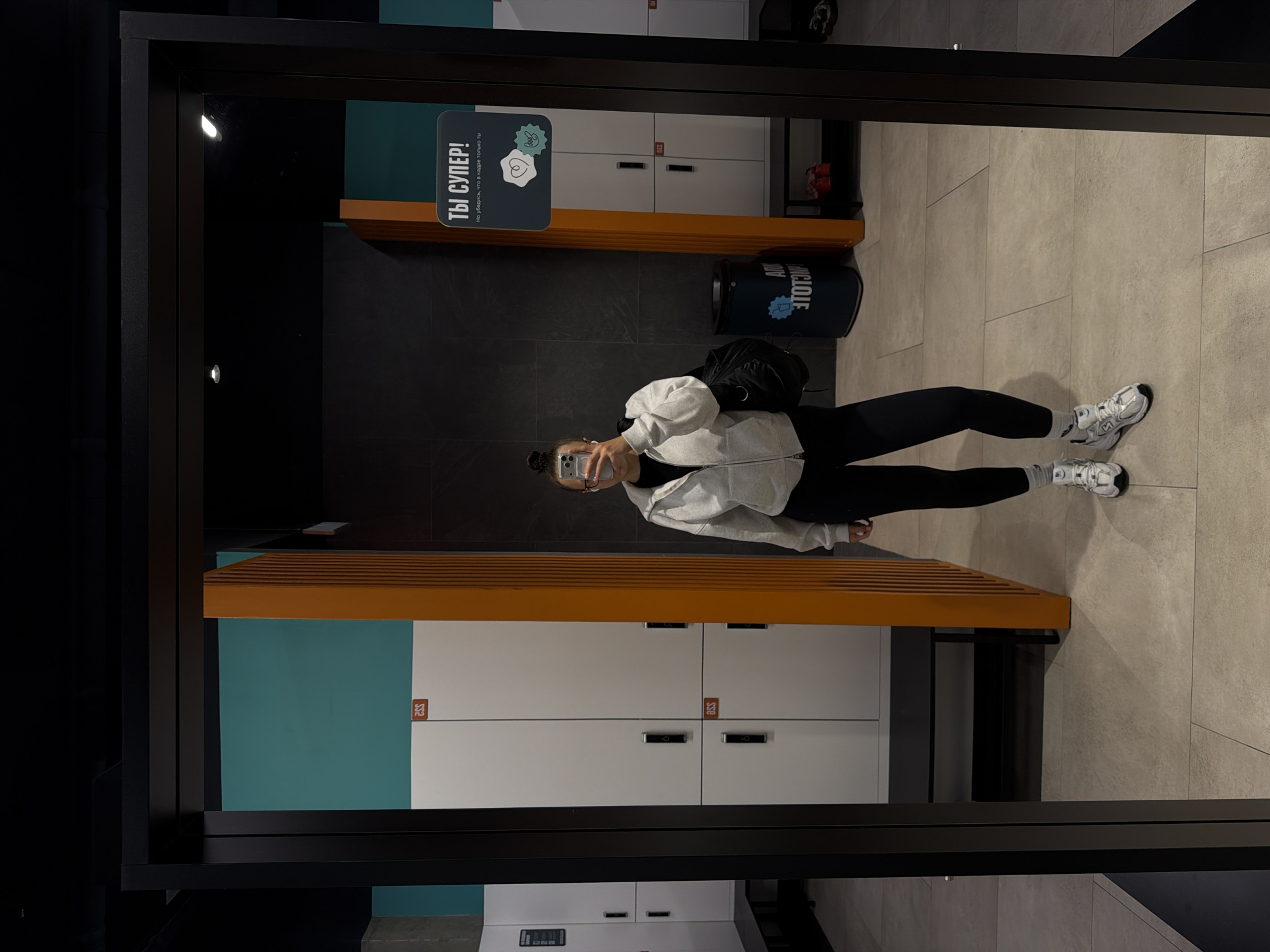 The image shows a person in a mirrored gym setting. The individual is wearing black leggings, a light-colored hoodie, and white sneakers. They are holding a mobile phone, presumably taking the picture in the mirror. A black backpack is slung over their shoulder. The environment includes lockers with orange accents and a sign with Cyrillic text "ТЫ СУПЕР!". A black trash can with Cyrillic text is also visible. The floor is tiled. There is no explicit indication of the city of Podolsk. The location is a gym changing room.