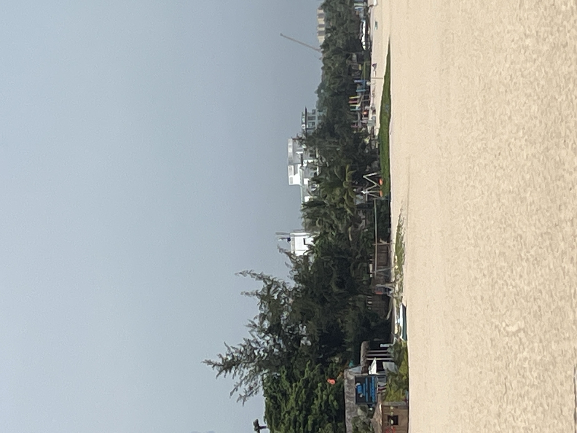 The image depicts a beach scene in Mũi Né, Vietnam, under an overcast sky. In the foreground is a vast expanse of pale sand, showing slight undulations. In the mid-ground, a line of lush green trees and vegetation separates the beach from a cluster of buildings and structures. Among these are several modern white buildings with multiple stories and balconies, suggesting hotels or resorts. To the left, a small, rustic wooden hut or kiosk is visible, with signage above it. Further to the right, playground equipment, including a swing set with orange seats and a colorful structure, is set up on the sand. In the distance, a construction crane is visible, hinting at ongoing development in the area. The overall impression is of a coastal resort town.