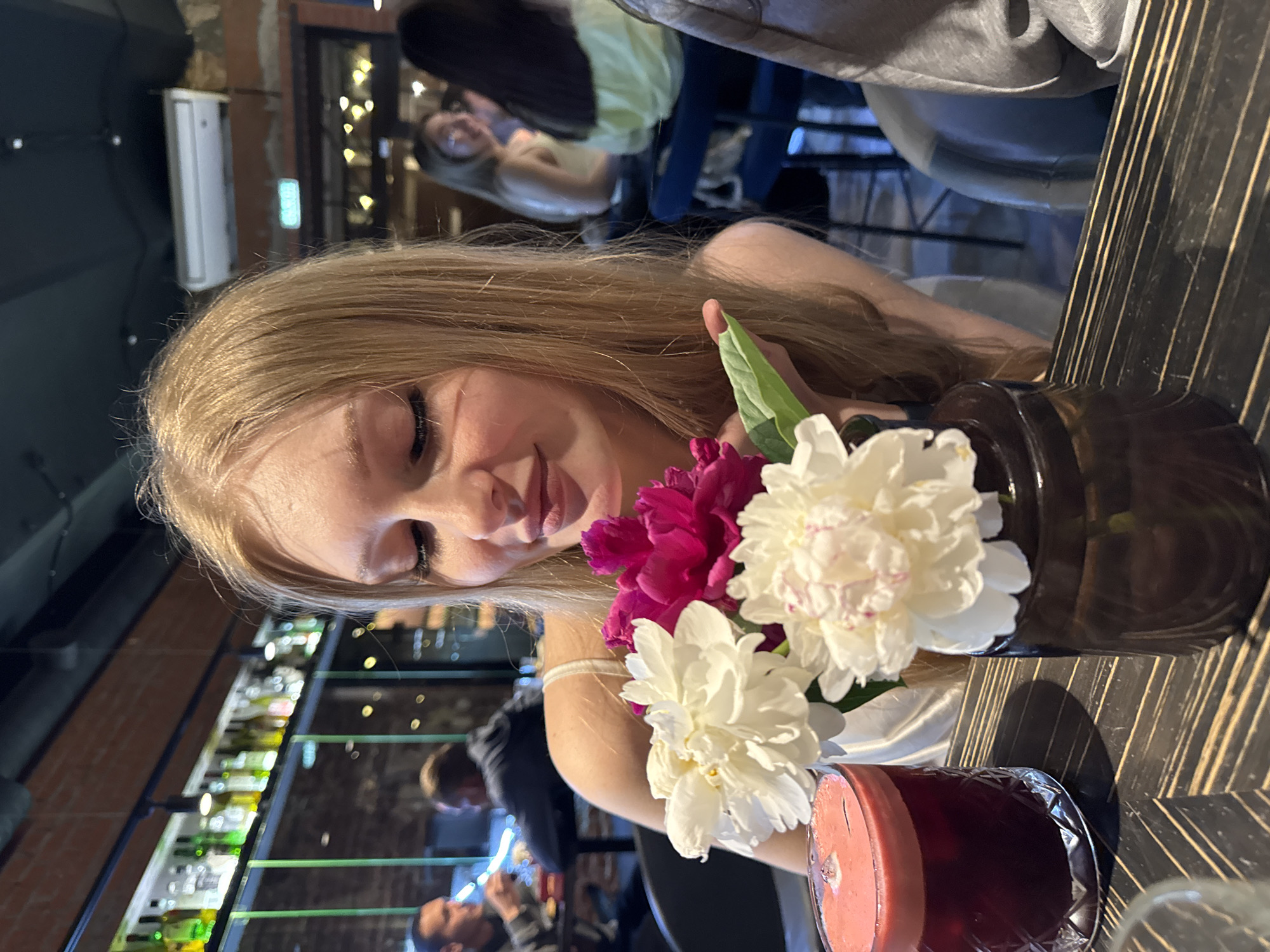 A young woman with blonde hair is seated at a table in a dimly lit establishment, possibly a bar or restaurant, with a bouquet of pink and white peonies in a dark brown vase in the foreground. A glass of dark red liquid, likely a drink, is also on the table to the left of the flowers. The woman is looking down towards the flowers with her eyes closed and a slight smile. In the background, other patrons are visible, and shelves lined with bottles can be seen behind a glass partition. The overall atmosphere suggests a relaxed evening setting. While the specific location in Perm, Russia, cannot be determined from the image alone, the presence of Russian signage in the background (a green exit sign) suggests it is indeed in Russia.
