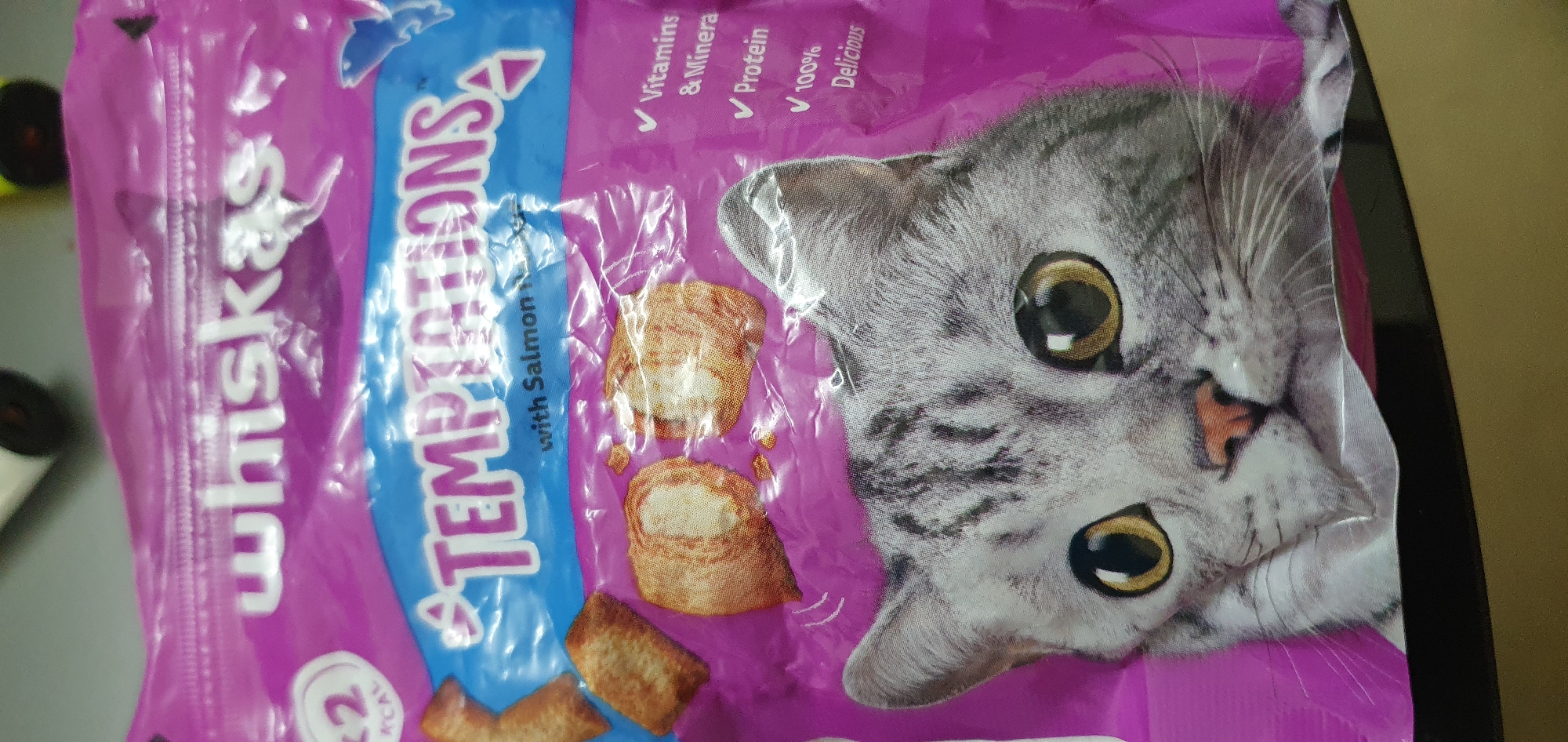This media file is a close-up, slightly angled photograph of a bag of Whiskas Temptations cat treats with salmon flavor. The bag is predominantly purple with blue and white lettering. The "Whiskas" brand name is visible vertically on the left side of the bag, followed by "Temptations" in a larger font. Underneath "Temptations" is text that appears to say "with Salmon".
To the right of the text, there are three visual representations of the cat treats themselves, appearing as small, golden-brown, bite-sized pieces. A prominent feature of the bag is a large, realistic image of a grey tabby cat's face, which takes up a significant portion of the right side of the frame. The cat's large, golden eyes are clearly visible, and its whiskers and nose are also depicted.
Above the cat's face and to the right of the "Temptations" text, several checkmarks are visible next to bullet points describing the treats: "Vitamins & Minera", "✓ Protein", and "✓ 100% Delicious". Near the bottom left of the bag, a circular graphic with the number "2" and the word "ACAL" is partially visible. The packaging appears to be made of a crinkled plastic material. The background is out of focus but suggests an indoor setting with some yellow and black objects on the left, and a plain, light-colored wall or surface on the right. There are no people visible in the image. The context provided indicates the location is Bat Yam, Israel.