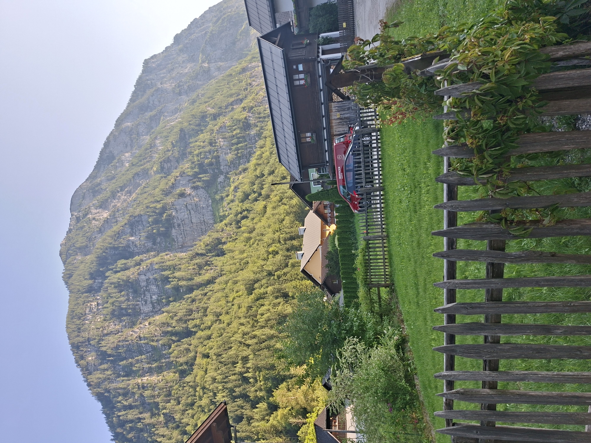 The image depicts a village nestled at the base of a steep, densely forested mountain in Hallstatt, Austria. A rustic wooden fence, weathered and showing signs of age, runs across the foreground, separating a meticulously manicured green lawn from a gravel path. Beyond the fence, several traditional Austrian houses with dark, sloped roofs are visible. One house, a prominent wooden structure, features multiple windows and decorative flower boxes. In front of this house, a red van with "HOFINGER" lettering and a dark sedan are parked. The mountain dominates the background, its rocky face partially obscured by a dense cover of evergreen trees, indicating a challenging terrain. The sky above is a clear, pale blue, suggesting fair weather and possibly early morning or late afternoon light due to the soft illumination. The overall scene conveys a sense of tranquil rural life in a stunning natural setting.