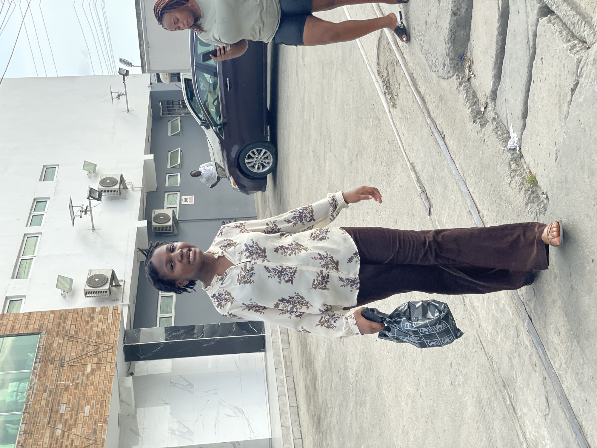The image, captured outdoors in Ado, Nigeria, shows a street scene under an overcast sky. In the foreground, a young Black woman with braided hair smiles and looks directly at the camera. She is wearing a white, long-sleeved blouse with a blue and brown floral pattern, paired with dark brown wide-leg trousers. In her left hand, she holds a crinkled black plastic bag featuring a white pattern of stylized cars and some indistinct text, including the number "13". Her light-colored sandal on her right foot is visible.
Behind her, to the right, another Black person with braided hair is partially visible, engrossed with their phone. They wear a light-colored t-shirt and dark shorts and stand next to a dark-colored sedan or small SUV. Further in the background, near the car, a Black man in a white shirt is also partially visible.
The setting includes multiple buildings. A prominent white, multi-story building features several windows (some with green frames), numerous external air conditioning units, and wall-mounted floodlights. Adjacent to it is a darker grey building with white-framed windows. A section with a brown brick facade and large glass windows, and a ground-level area with white and black marble-like tiling, also appear. The ground is a paved concrete surface with some cracks and an embedded metal strip. The casual poses of the subjects suggest a relaxed daytime atmosphere in an urban or semi-urban environment.