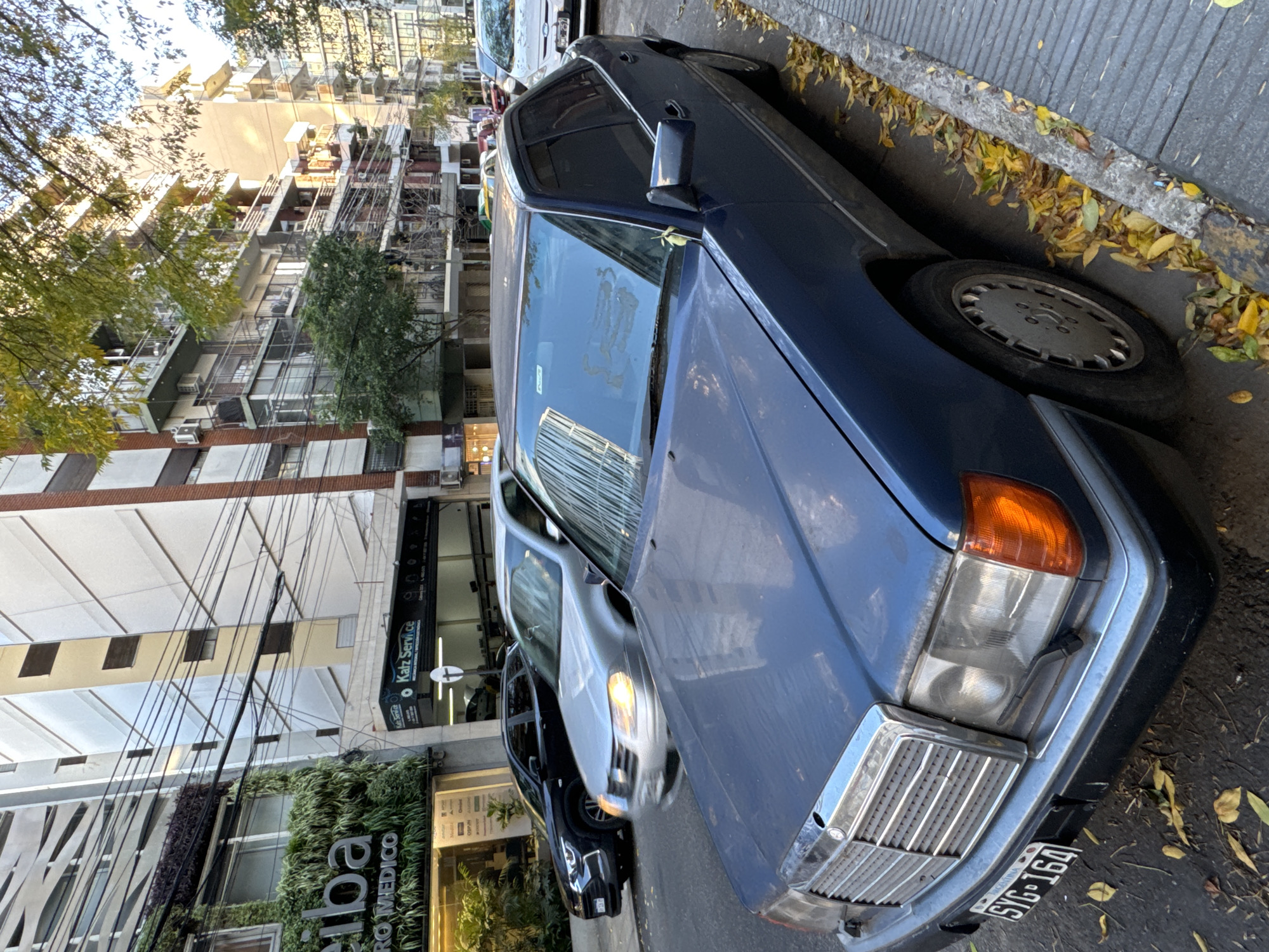 A blue Mercedes-Benz car is parked on a city street, next to a curb covered with fallen leaves. The license plate reads "SIG-164". Behind the Mercedes, other cars are parked. Buildings line the street, including one with vertical greenery and the word "medico" visible. The architecture and signage suggest this is in Buenos Aires, Argentina. Overhead power lines are visible.