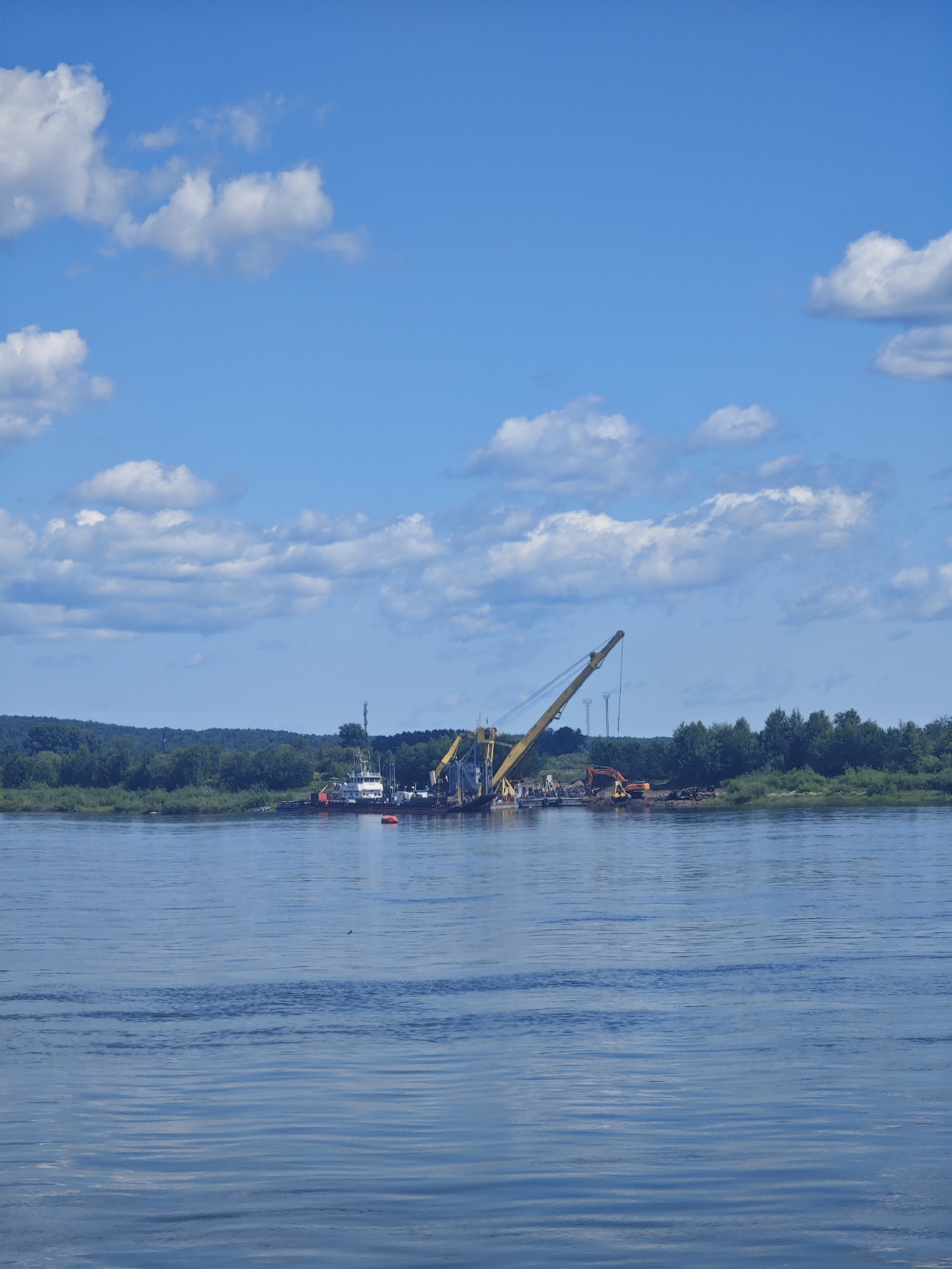 This is a photograph of a wide, calm river with a line of trees and vegetation forming a dense forest on the distant bank. The sky above is a vibrant blue with scattered, fluffy white clouds. In the middle of the river, near the shore, is a work barge equipped with a large yellow crane. Adjacent to the barge, another smaller vessel and what appears to be construction equipment are visible. The scene suggests ongoing work or a construction project taking place on the river. The location is Strelka, Russia.