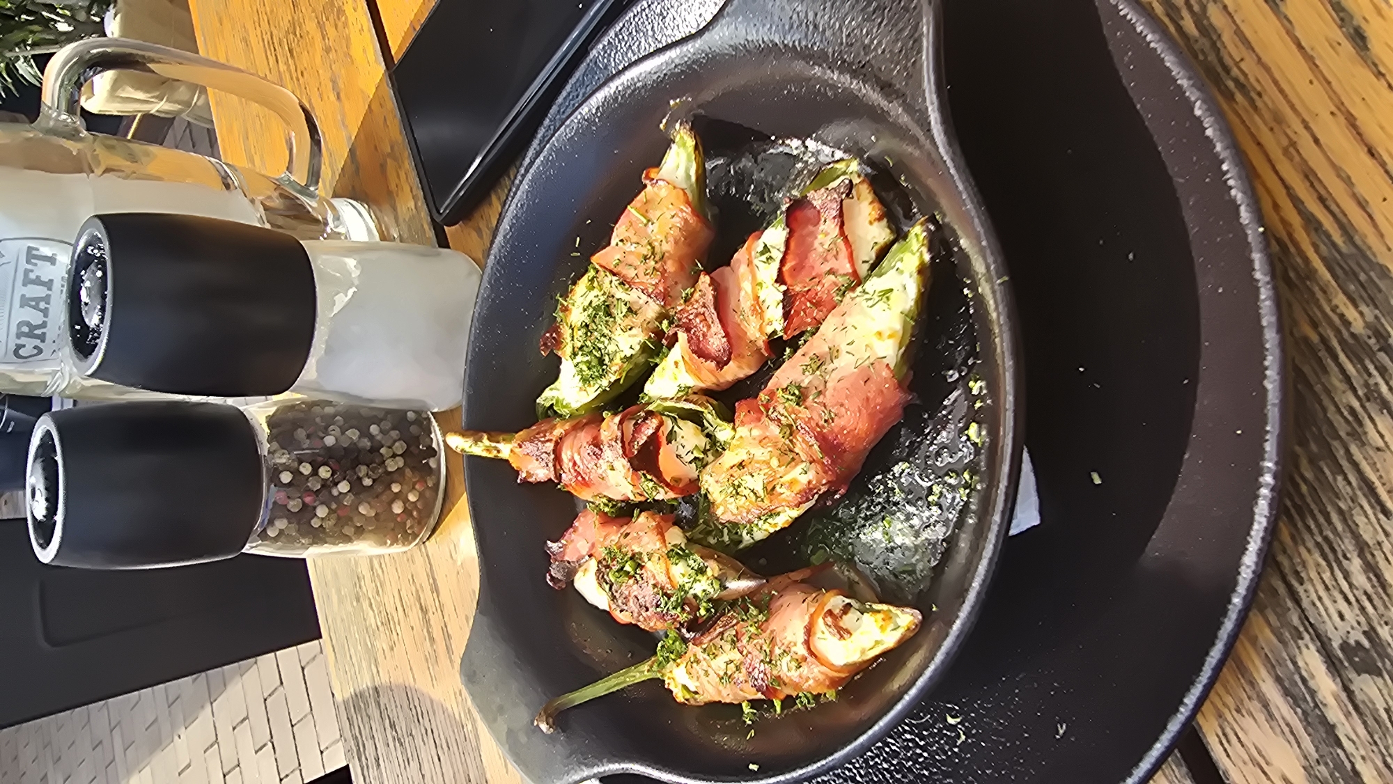 An outdoor table at a restaurant in Varna, Bulgaria, displays a dish of jalapeño poppers wrapped in bacon, sprinkled with herbs. The poppers are served in a black, oval cast-iron skillet. In the background, a glass mug with "CRAFT" printed on it, and a salt and pepper grinder set are visible on the table. The table is made of weathered wood planks. Sunlight illuminates the scene, casting shadows.