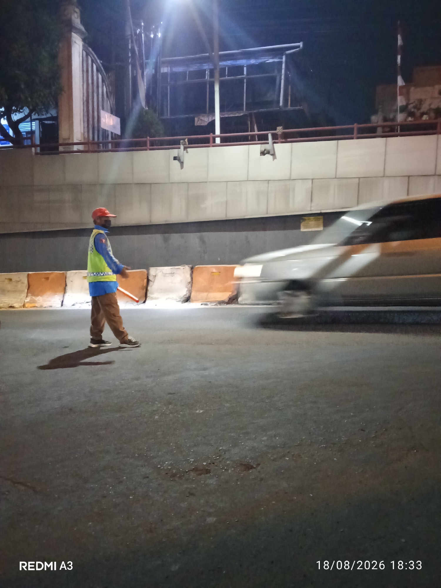 In the city of Purbayan, Indonesia, a traffic controller stands on a road at night, illuminated by overhead lights. The controller, wearing a red cap, a blue shirt, a high-visibility yellow vest with reflective stripes, and brown pants, is positioned in the left half of the frame. They are holding a reflective baton in their right hand, and their left hand is extended. The road surface is asphalt, with scattered debris. Orange and white concrete barriers are visible in the background, separating the traffic lanes. A silver car is in motion to the right of the controller, blurred due to its speed. Overhead, a structure with metal beams and a blue tarp is present, suggesting ongoing construction or maintenance. The scene is captured with the timestamp "18/08/2026 18:33" and the device indicator "REDMI A3" at the bottom of the image.
