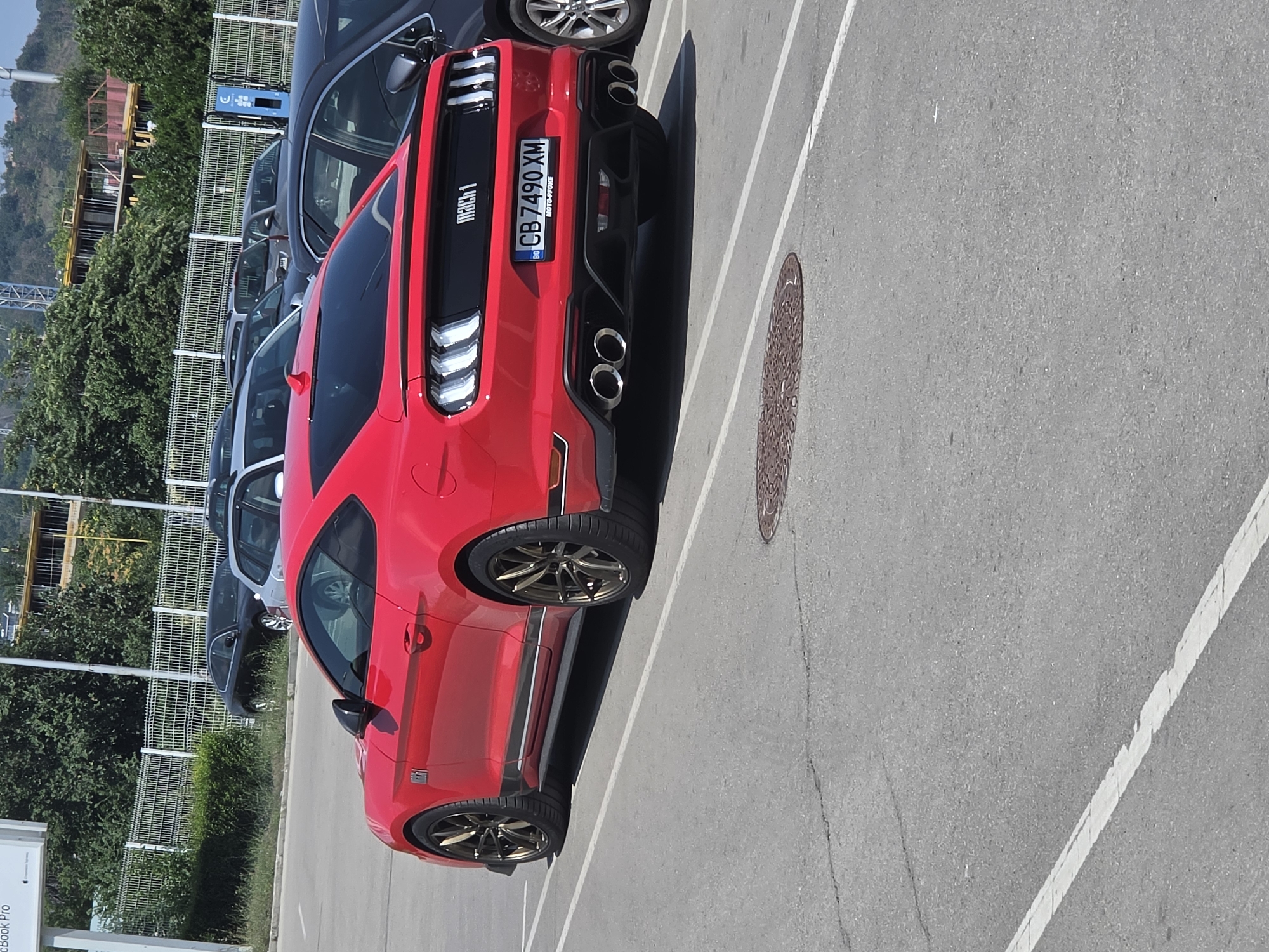 The image shows a red Ford Mustang Mach 1 parked in an outdoor asphalt parking lot. The car is positioned diagonally, with its rear facing the camera. It has a license plate that reads "CB 7490 XM BG". Other cars are visible in the background, including black sedans and a silver hatchback. Behind the cars, there is a metal fence, green bushes, and trees. In the far background, buildings under construction are visible. The scene appears to be a sunny day. Based on the license plate format, this scene is located in Varna, Bulgaria.