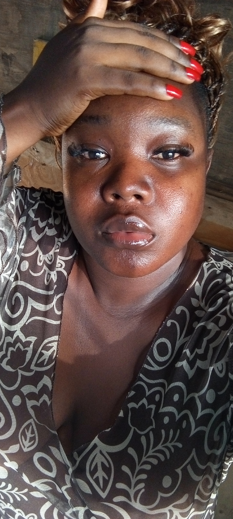 A dark-skinned woman is positioned in the center of the image with her hand on her forehead and red nail polish on her nails. She is wearing a patterned brown and white top. Her makeup includes false eyelashes. The location cannot be determined to be in Lagos, Nigeria without further contextual clues.