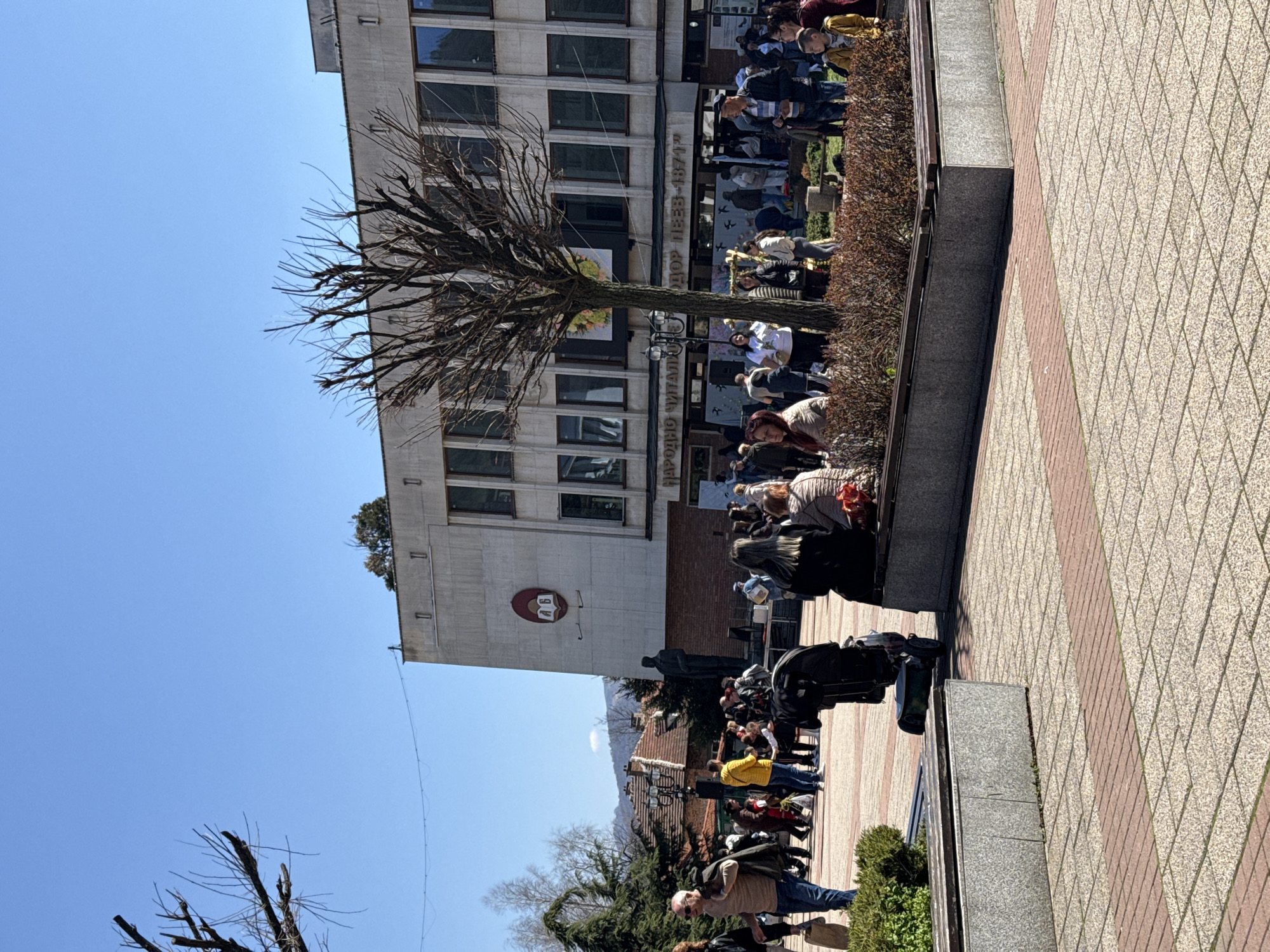 The image shows a group of people gathered in front of a building in Etropole, Bulgaria. The building has a sign in Cyrillic script that says "Народно Читалище 'Тодор Пеев-1871'." There is a bare tree in the foreground. A statue and some mountains are visible in the background. The sky is blue and mostly clear. The people seem to be participating in an event. The ground is paved with patterned stones.