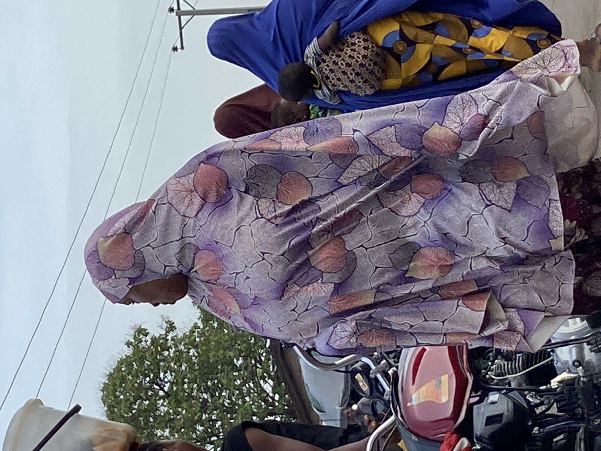 The image captures an outdoor scene, likely in Mararaba, Nigeria, featuring several individuals and a motorcycle.
In the center-right, a person is prominently visible, wearing a light purple head covering and a garment adorned with distinctive leaf-like patterns in various shades of purple, pink, and gray. Their profile is seen, looking towards the top of the frame.
Further towards the top-left, another individual is present, clad in a vibrant blue head covering and a garment with a striking yellow and blue geometric design. A small child, wearing a striped top, is held close by this person. Parts of a third person, dressed in reddish-brown, are partially visible behind them.
The lower-right and central sections of the image are dominated by a red motorcycle, showcasing its gas tank, engine parts, and chrome accents. In the lower-left corner, a white plastic container or bucket is partially visible.
The background (upper-left) features a bright, overcast sky with utility poles and overhead power lines stretching across it. Green tree foliage is scattered towards the left and lower parts of the frame, providing a natural element to the setting.
The scene depicts a moment of daily life, possibly people traveling or waiting alongside a road, with the motorcycle suggesting a mode of transport. The time of day appears to be daytime due to the bright ambient light. No specific activity is clearly underway, and no discernible text is visible anywhere in the image. Emotions of the individuals cannot be determined from their obscured or distant visages.