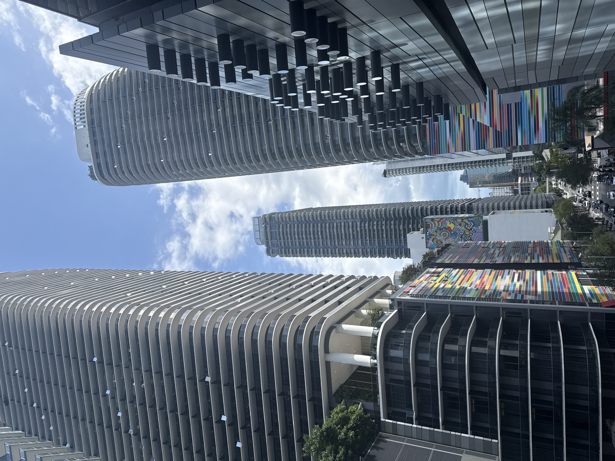 The image showcases a view of the cityscape in Miami, United States. The angle is from below, looking upwards towards the sky, revealing several skyscrapers. The architecture is modern and varied, with a focus on verticality and curvilinear forms. One building features a unique design with continuous horizontal ribs spanning the facade. Another structure displays an exterior adorned with a colorful, striped pattern that seems to wrap around its sides. Clear skies with scattered clouds are visible between and above the buildings, emphasizing the height of the structures. The scene is bathed in daylight, highlighting the textures and contrasts of the urban environment.