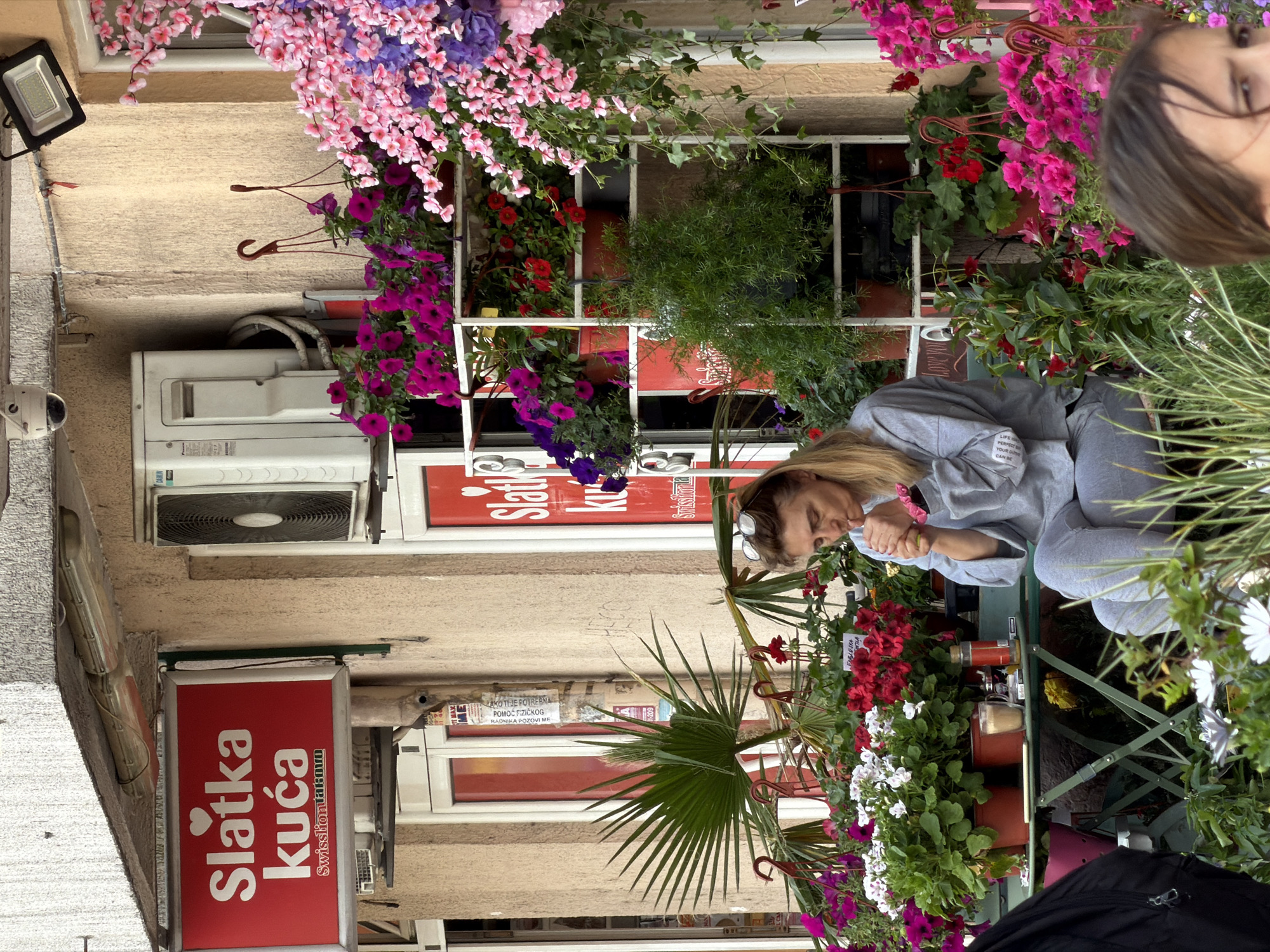 The image shows a street scene in Budva, Montenegro. A woman in a grey sweatsuit is looking at something on a small green metal table surrounded by potted plants with red and white flowers. Behind the table, there is a store with a sign that reads "Slatka Kuca Swissliontakovo". The building has an air conditioning unit and a security camera. The storefront is decorated with flowers in various colors. There is a lamppost and a black bag partially visible at the bottom.