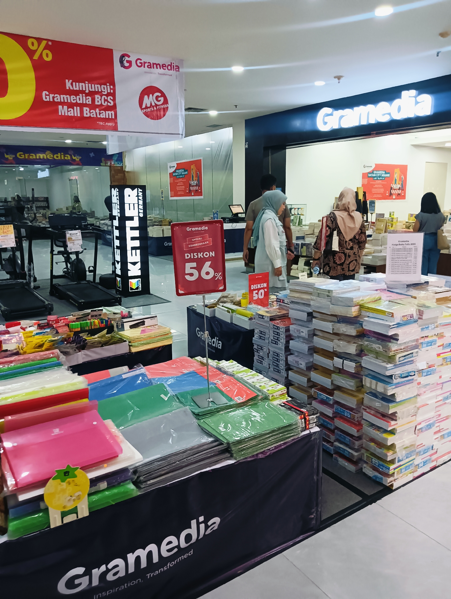 The image depicts the interior of a Gramedia bookstore, a well-known retail chain in Indonesia, likely located within the BCS Mall in Batam City. The scene is filled with merchandise, predominantly books and stationery. In the foreground, a black display cloth prominently features the Gramedia logo and the tagline "Inspiration, Transformed." On top of this cloth, stacks of colorful folders and stationery items are neatly arranged. To the right, tall stacks of books are visible, some with price tags and discount information, including a red sign indicating "DISKON 56%" (56% Discount) and another with "DISKON 50%" (50% Discount).
In the middle ground, a display of KETTLE sports equipment, including treadmills and exercise bikes, is set up. Further back, customers are browsing, with one person wearing a hijab and another in the background looking at books. A large Gramedia sign with illuminated lettering is visible on a dark blue backdrop above the store entrance. In the upper left corner, a promotional banner with a large "%" symbol and text in red and white reads "Kunjungi: Gramedia BCS Mall Batam" (Visit: Gramedia BCS Mall Batam). The environment appears to be a typical retail space with bright overhead lighting.