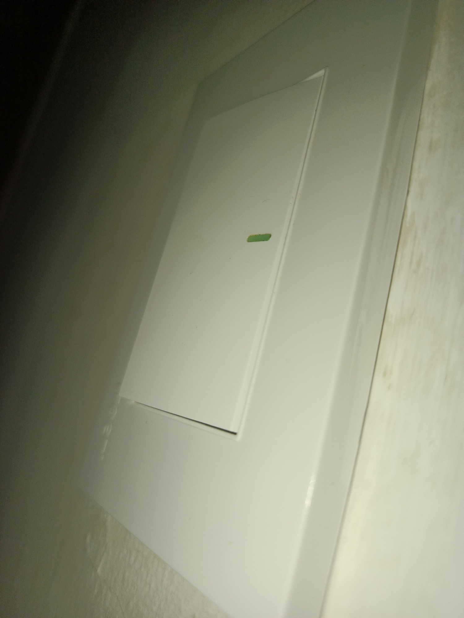 A close-up, angled shot focuses on a white, rectangular light switch installed on a wall. The switch appears to be in the "off" position, with a small green sticker placed near its center. The wall itself has a mottled, slightly discolored appearance. The lighting is dim, casting shadows that add depth to the image. No people or other objects are visible. The location in Los Robles, Venezuela is not verifiable from the image.