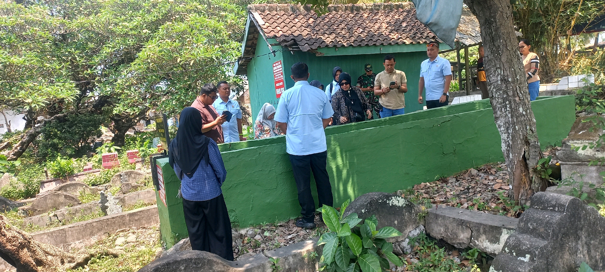 The image depicts a group of people gathered at a cemetery in Bandar Lampung, Indonesia. The cemetery appears to be located on a hillside with numerous gravestones, many of which are partially overgrown with vegetation. A prominent green-painted wall or structure runs across the foreground, separating the group from some of the graves.
In the center of the image, several individuals are interacting. One person, wearing a light blue shirt, is facing away from the camera, appearing to look at something on the green wall. To their left, a man in a brown shirt is holding a tablet or phone, possibly showing it to another person. A woman in a black hijab and patterned top is also looking in the same direction. Further to the left, a woman in a blue patterned top and black hijab stands with her back to the camera.
In the background, a small green building with a tiled roof is visible. There are also other people present, including someone in a military uniform. The environment is outdoors, under a bright sky, with trees and foliage surrounding the cemetery. The overall scene suggests a visit or inspection of the cemetery.