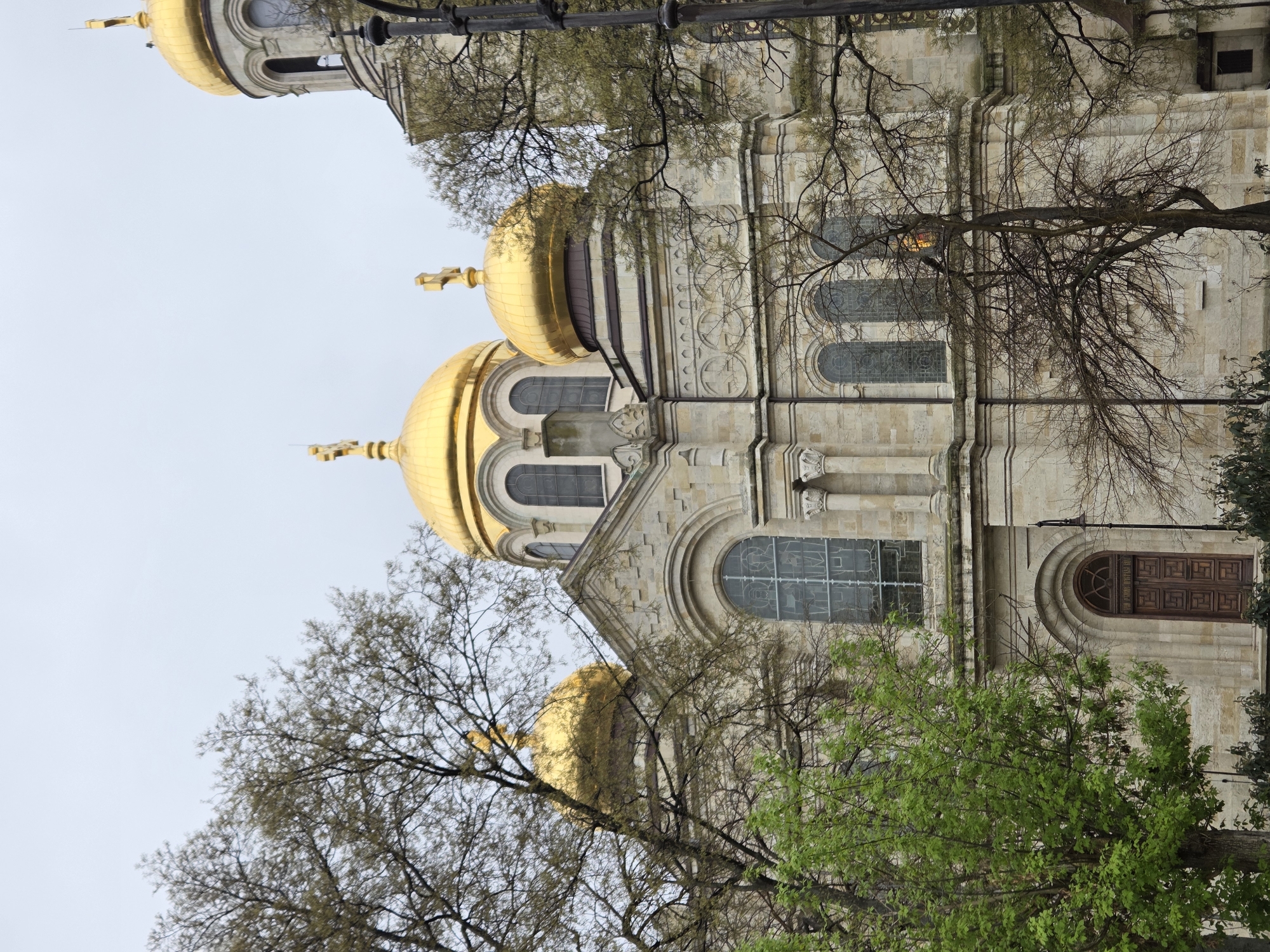 This low-angle, outdoor shot captures the ornate facade of a grand Orthodox cathedral, likely the Dormition of the Theotokos Cathedral in Varna, Bulgaria. The building is constructed of light-colored stone, featuring intricate architectural details such as decorative pilasters, cornices, and multiple arched windows.
Several prominent golden domes, topped with crosses, are visible, reflecting the ambient light. One dome belongs to a taller bell tower or central cupola, while others are lower on the structure. The windows display patterned or stained glass. On the right side of the main image, a large, dark wooden door with a rounded top and geometric paneling is partially visible. Above this door, within a semi-circular arch, there is an inscription, though the Cyrillic text is illegible due to resolution.
The cathedral is partially obscured and framed by trees. Some trees in the foreground have bare, intertwining branches, while others show the fresh green foliage of spring, indicating the time of year. The sky is bright and overcast, suggesting daytime with diffused light and no direct sunlight. No people or specific activities are depicted; the image focuses solely on the architecture and its natural surroundings.