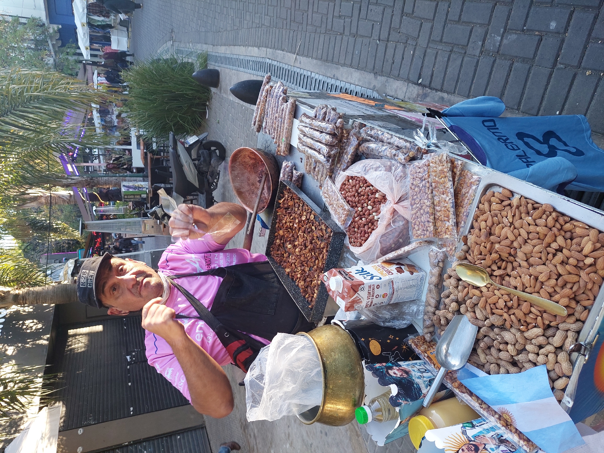 The image shows a man in a pink shirt and black apron standing behind a vendor's table laden with nuts and other items, likely in Buenos Aires, Argentina. The man is giving a thumbs-up gesture. On the table, there are various types of nuts, including peanuts, almonds, and candied nuts, arranged in trays and plastic packaging. Other objects on the table include a metal bowl, a carton of chocolate almond milk, a small Argentinian flag, and serving utensils like spoons and scoops. The background suggests an outdoor urban setting, potentially a pedestrian street with shops and restaurants. There is a palm tree to the left of the frame.