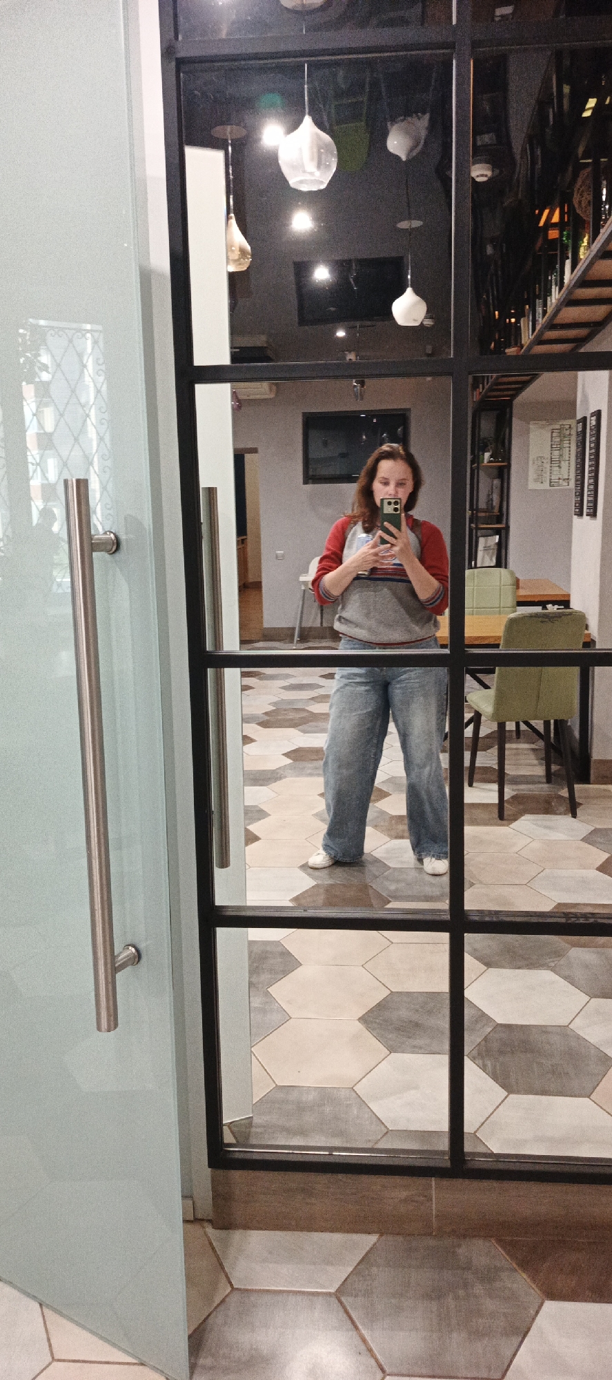 A young woman is taking a mirror selfie in what appears to be a cafe or restaurant. She is wearing a grey sweatshirt with red sleeves and blue jeans. The cafe has a modern design with hexagonal floor tiles and industrial-style light fixtures. In the background, there are tables and chairs, shelves with bottles, and a television mounted on the wall. The woman is holding a smartphone in her right hand, and her left hand is near her chest. The overall scene is well-lit.