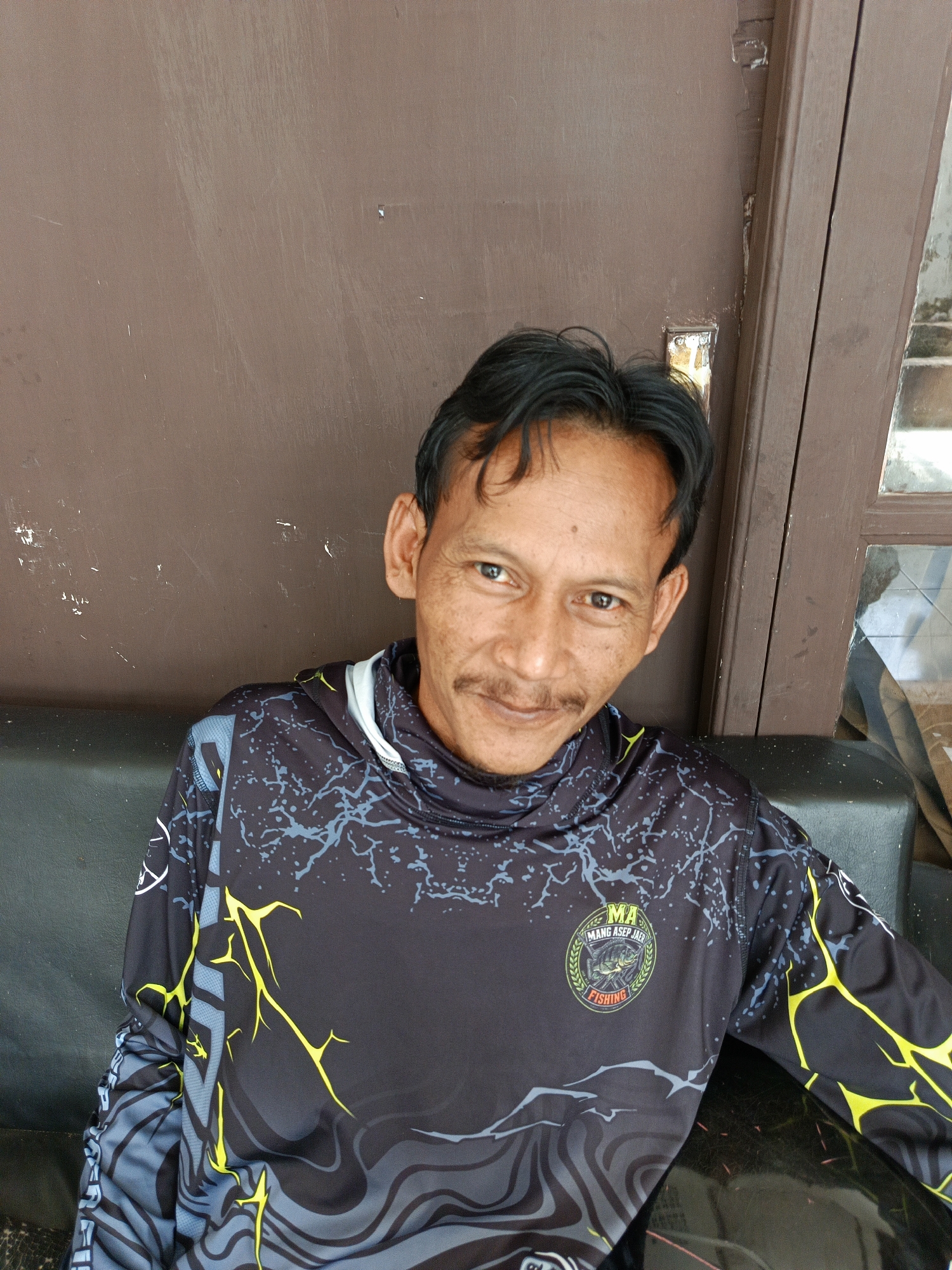A man is seated on a dark, possibly leather, couch, facing the camera. He has dark hair, a mustache, and appears to be middle-aged. He is wearing a black long-sleeved shirt with a complex design featuring yellow lightning-like patterns and gray lines. A circular logo is visible on the left side of his chest, which reads "MA MANG ASEP JAEF FISHING". Behind him is a dark brown door or wall with some visible markings and a section of a window with reflections. The man is positioned in what appears to be an outdoor or semi-outdoor setting, possibly near a doorway in Margajaya, Indonesia, given the context provided.