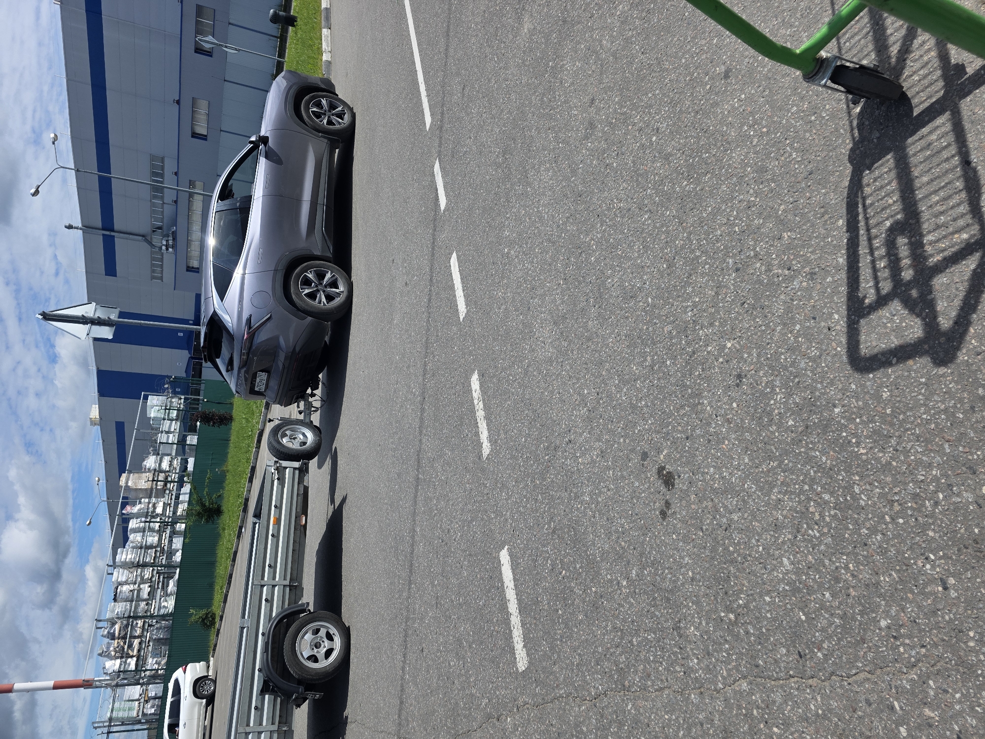 A gray SUV is parked on the right side of a paved road, towing a small utility trailer. The SUV is facing away from the camera, with its rear bumper and wheels visible. The trailer is attached to the SUV via a hitch and has two wheels on its left side, one of which is prominently displayed. The road is asphalt and appears to have white dashed lane markings. In the background, a large industrial-looking building with a blue and gray facade stands under a partly cloudy sky. Green fencing and some vegetation are visible in front of the building. A white SUV is parked on the left side of the road, further away from the camera. The overall scene suggests an industrial or commercial area on a clear day.
