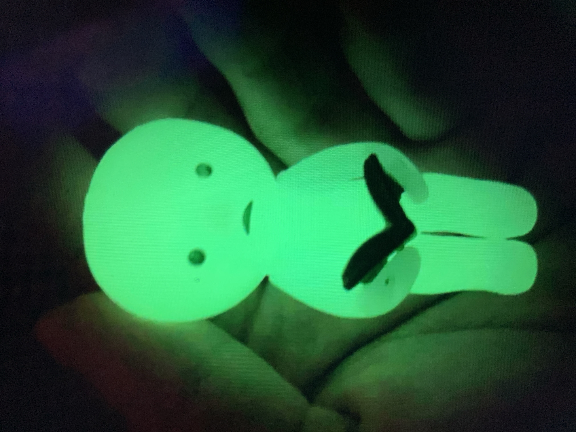 A glowing green, doll-like figure with a rounded head, small dot-like eyes, and a downturned mouth is held in two hands. The figure has a simplified humanoid form and is holding a dark object, possibly a book or tablet, with both hands at chest level. The background is dark and indistinct, with hints of reddish-brown and green tones, possibly from clothing or the environment. The image is taken in close-up, focusing on the figure and the hands holding it. The location in Lyman, Ukraine, is not determinable from the visual information presented in the image.