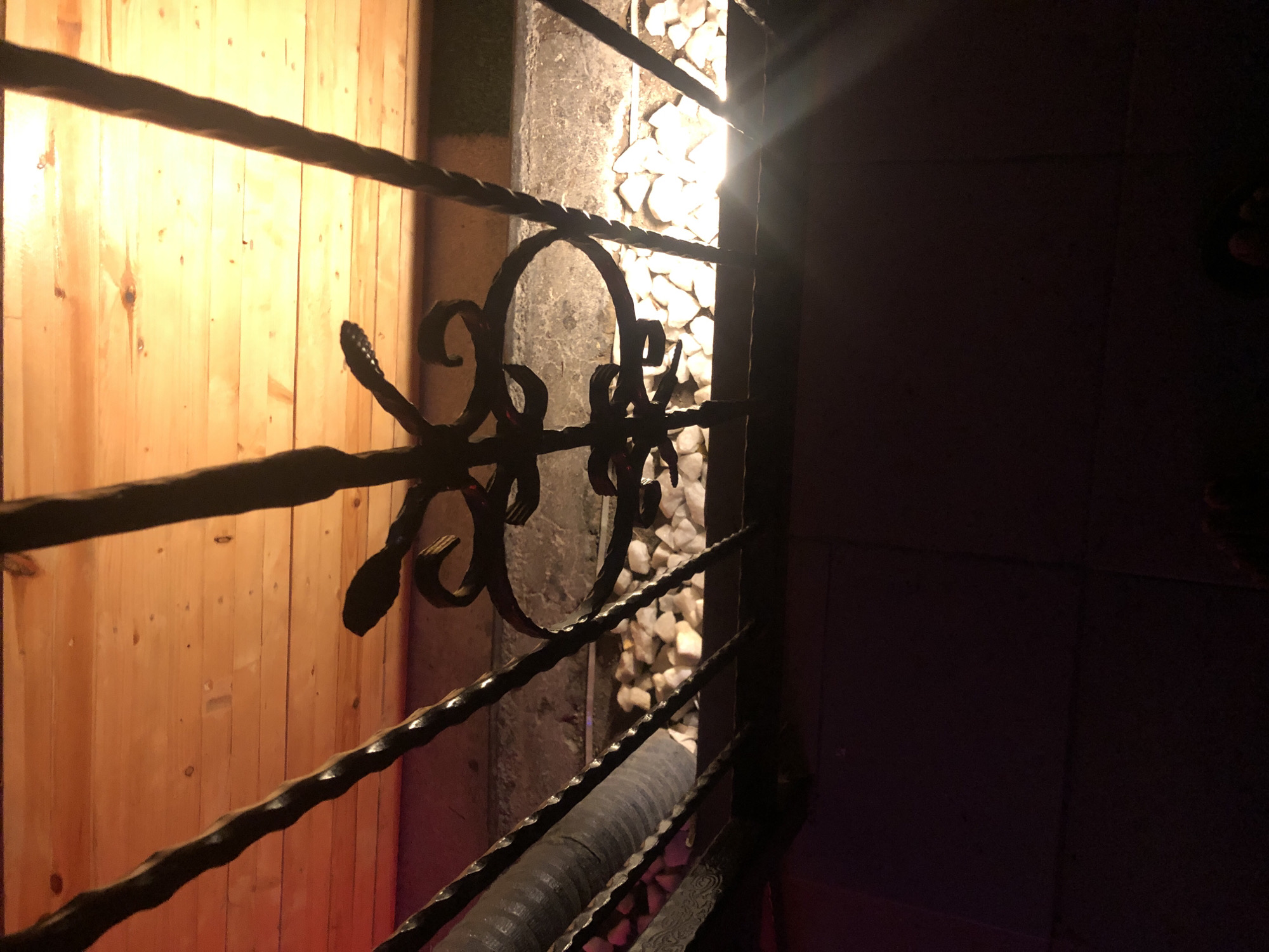 The image depicts a close-up view of decorative wrought iron bars, possibly part of a fence or gate, set against a background of wooden planks. Below the bars, there is a layer of white rocks illuminated by a bright light source, creating a strong glare. The overall lighting suggests an outdoor setting, possibly at night or in a dimly lit area, with warm-toned illumination from the wooden planks and the bright light on the rocks. The lower portion of the image is dark, showing what appears to be a tiled or paved surface. There are no people or specific identifiable landmarks of Howayi, Nigeria present in the image.