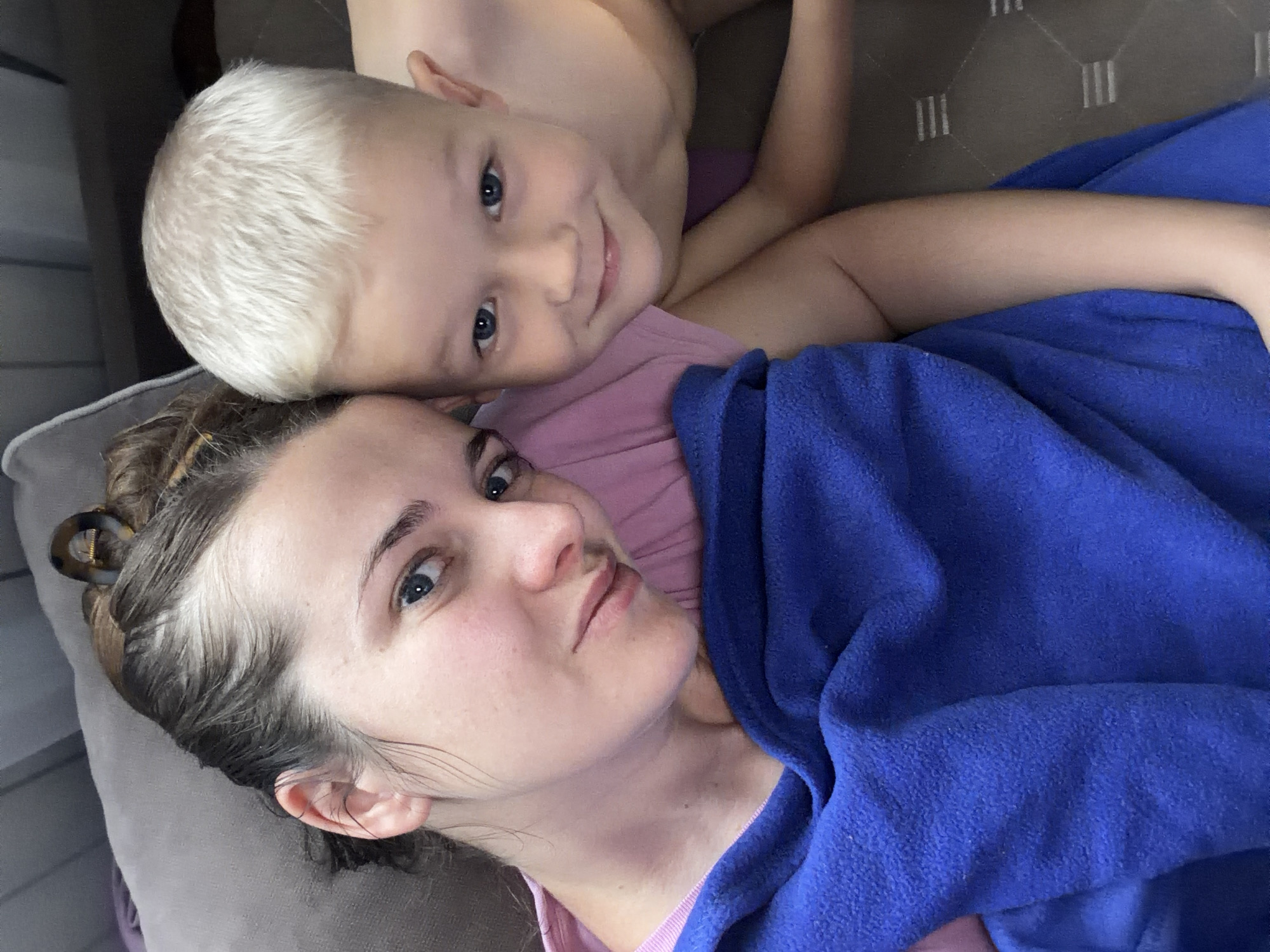 The image captures an intimate, overhead perspective of a woman and a young child, likely a mother and her son, relaxing together indoors in Sochi, Russia. Both individuals are lying down, looking towards the camera, suggesting a selfie or a closely taken portrait.
The woman, in the foreground and lower half of the frame, has dark hair, some strands appearing lighter, pulled back with a brown and clear hair clip. She has light skin, blue eyes, and a neutral, attentive expression. She is wearing a pink top, and a vibrant blue fleece blanket or towel is draped over her, covering her right arm and torso.
Above and slightly behind her, the child, with very short, light blonde or platinum hair and bright blue eyes, rests his head on her shoulder or chest area. He looks at the camera with a sweet, gentle smile, conveying contentment. He appears to be wearing a light-colored top, possibly pink. One of his arms is visible, resting near the woman's head.
The background features soft, textured gray upholstery, indicative of a sofa or bed, reinforcing the cozy, relaxed, indoor setting. The lighting suggests it is daytime. No text is visible in the image. The scene evokes feelings of warmth, affection, and peaceful family togetherness.