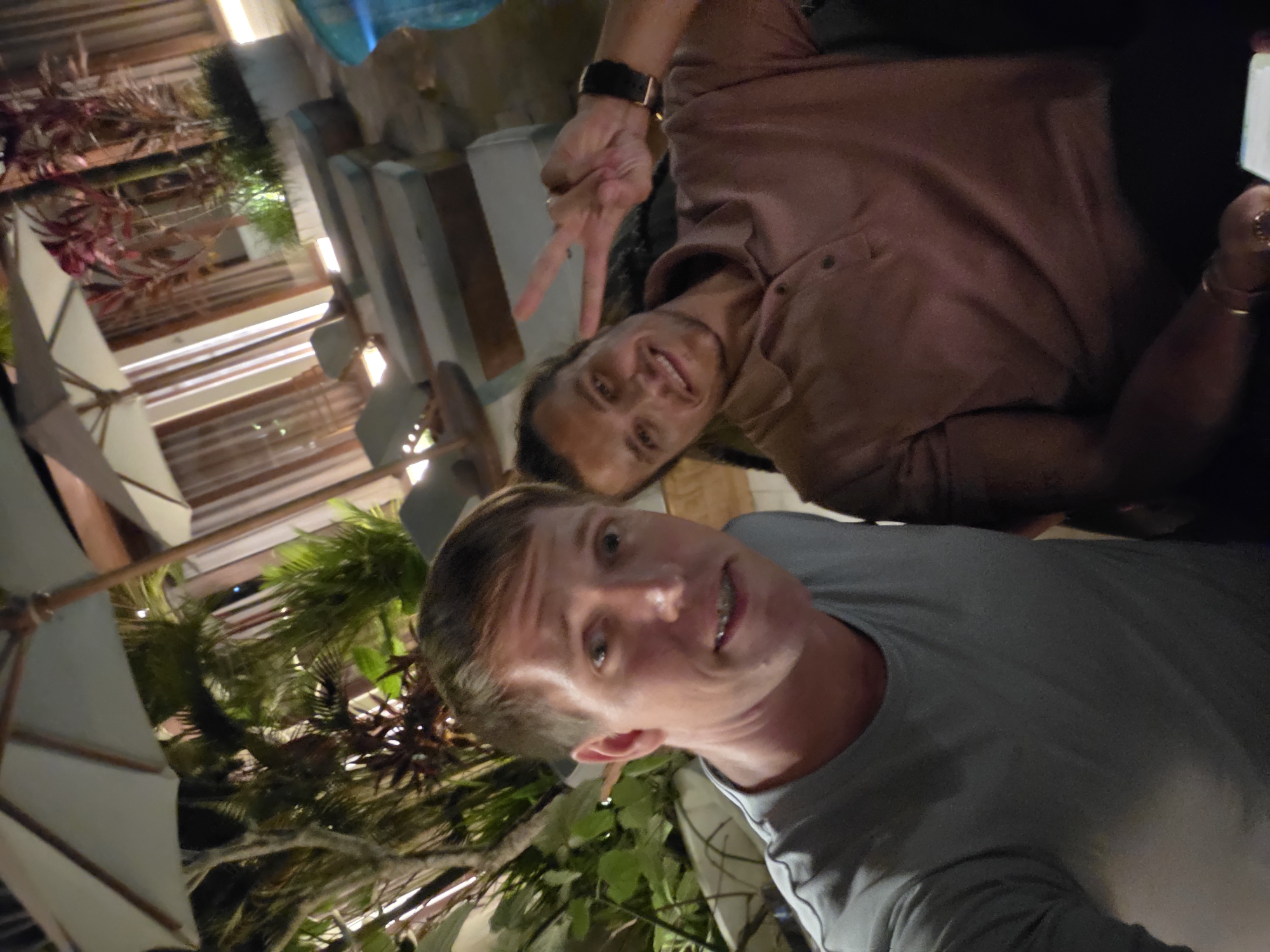 Two men pose for a selfie at a restaurant in the Ubud District, Indonesia. The man in the foreground wears a gray t-shirt and smiles. The man in the background wears a brown shirt and makes a peace sign. The restaurant is decorated with plants and umbrellas.