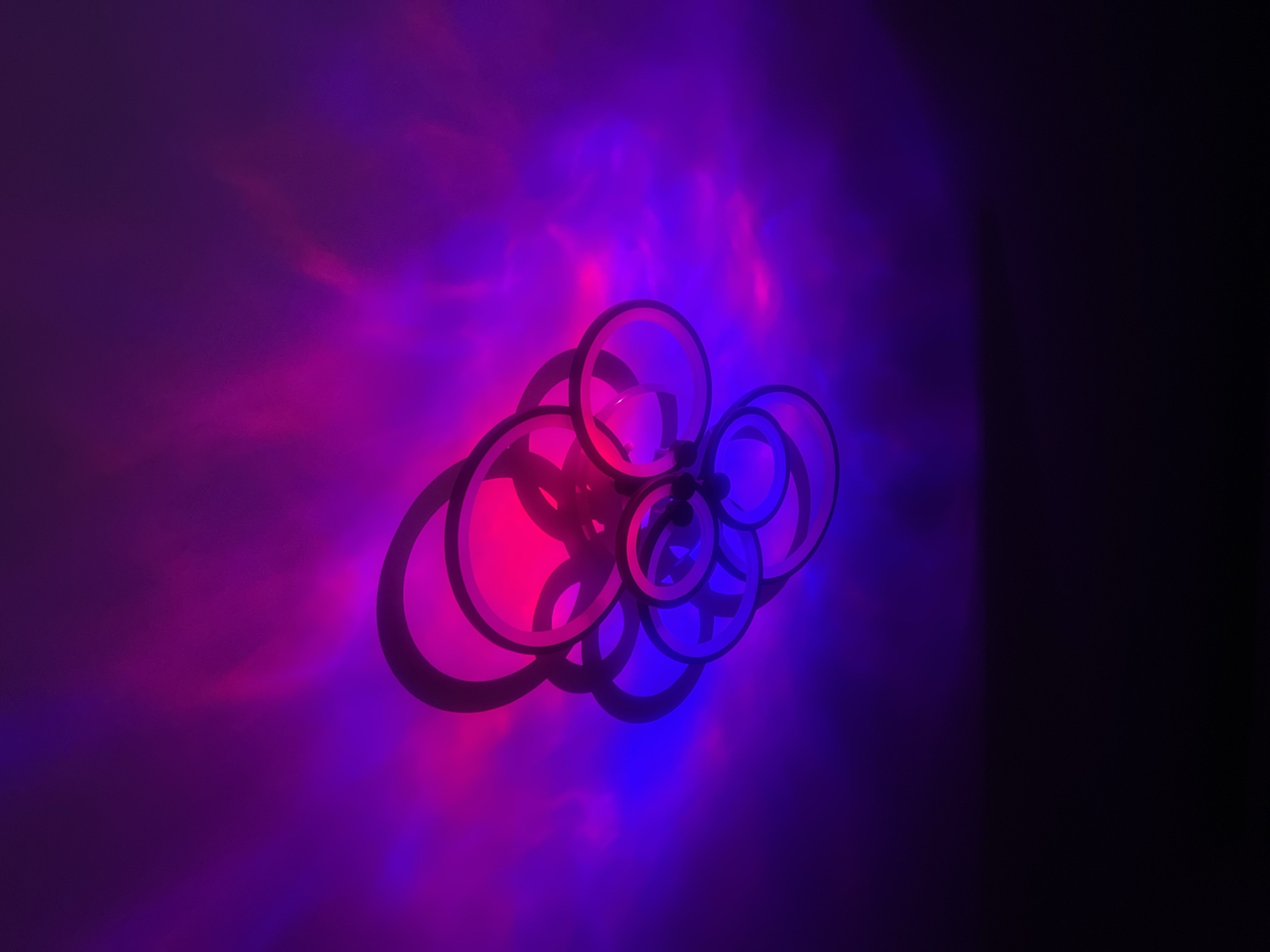 The image features a cluster of interlocking black circles against a background gradient of purple, pink, and blue light. The circles cast dark shadows on the illuminated surface. The right side of the image fades into darkness. There is no information about the location or the city of Chaikovsky, Russia, provided. No people, subjects, actions, or interactions are depicted in the image.
