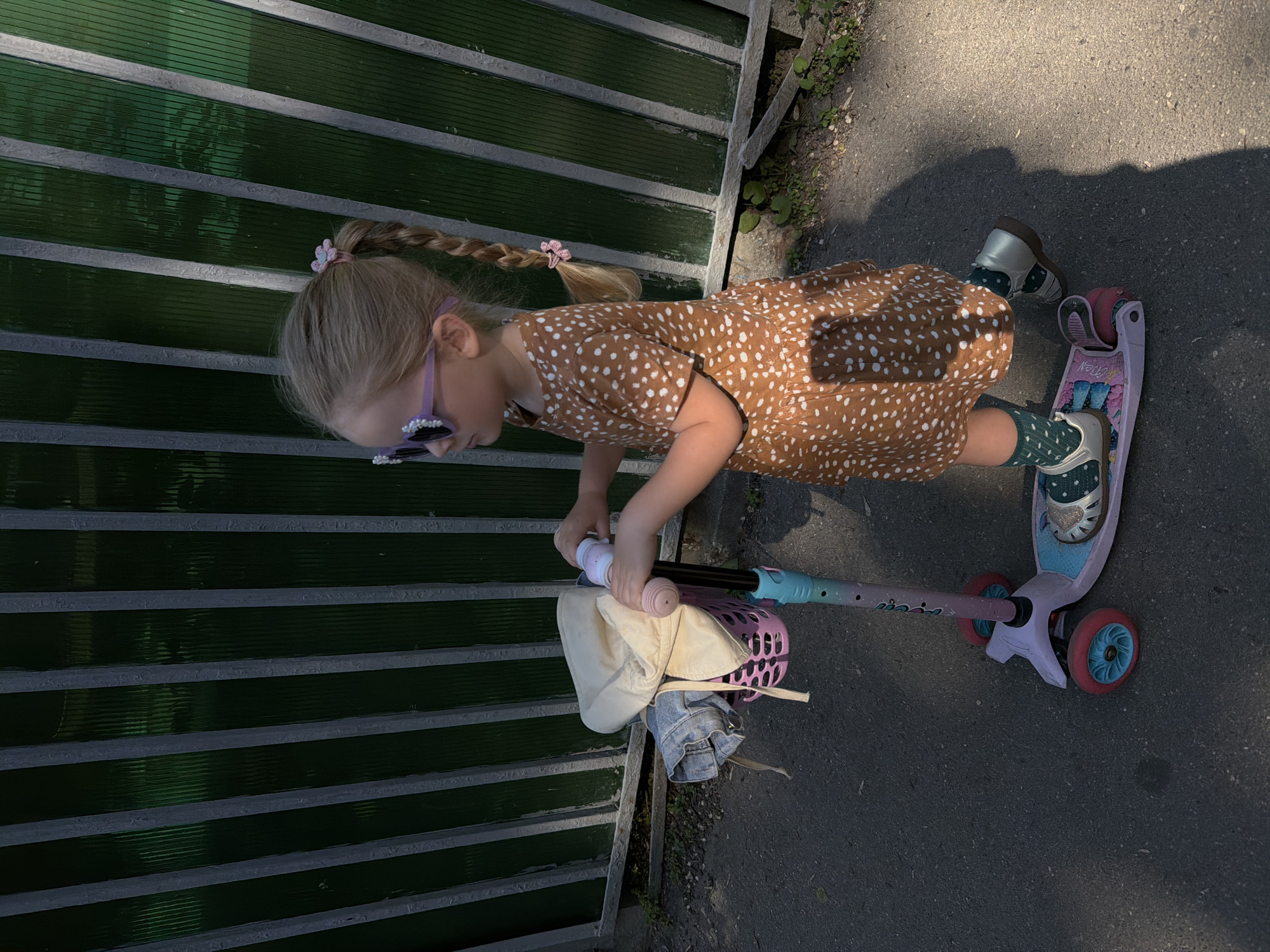 A young girl is riding a pink scooter on a paved surface. She is wearing a brown dress with white spots, green socks with white dots, and silver shoes. She has blonde hair in braids with pink hair ties. She is also wearing purple sunglasses. The scooter has a pink basket attached to the front handlebars which holds a beige hat and a piece of blue clothing. The girl is next to a green fence with dark horizontal bars. The location is Moscow, Russia.