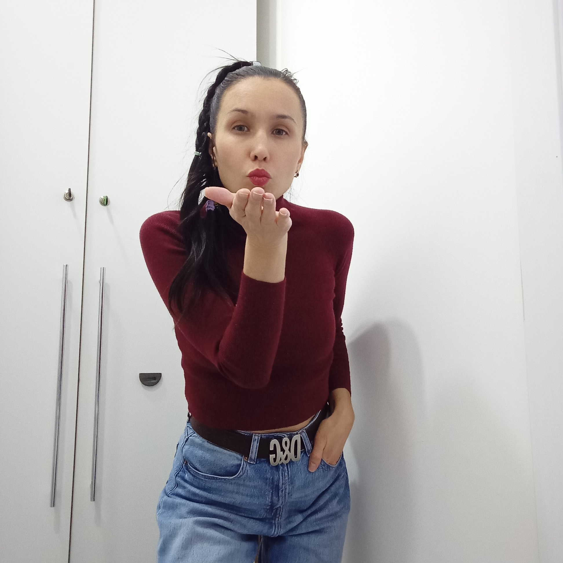 A young woman is captured in a mid-shot, posing indoors against a plain white background that appears to be a wardrobe or cabinet with silver handles and small locks. She is looking directly at the viewer and blowing a kiss, with her lips pursed and her right hand cupped near her mouth. Her expression is playful and affectionate.
She has long, dark hair, styled partially in a high ponytail with some braids or twists, and some loose strands fall over her shoulder. Small, colorful hair accessories are visible in her hair. She is wearing a dark red, long-sleeved, high-neck top that is cropped, revealing a sliver of skin above her light blue denim jeans. A prominent dark brown belt with a large, silver-colored buckle spelling "D&G" is cinched around her waist. Her left hand is casually tucked into the front pocket of her jeans.
The lighting is bright and even, consistent with an indoor setting, and no specific time of day is discernible. The overall scene suggests a casual, personal moment. The location context provided is Izhevsk, Russia.