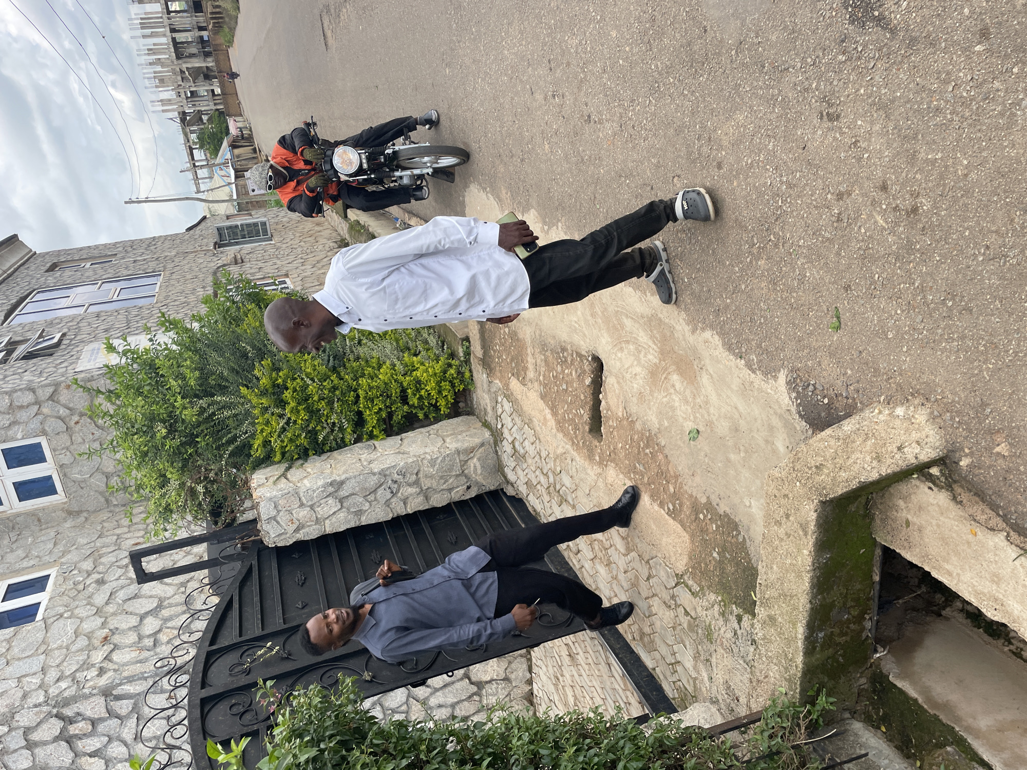 The image depicts a street scene in Gura Top, Nigeria. Two men are the primary subjects. The man on the left, wearing a dark blue long-sleeved shirt and black pants, stands near a large, ornate black gate and a stone-clad building. He is looking towards the man on the right and appears to be holding a cigarette. The man on the right, dressed in a white long-sleeved shirt and black pants with grey crocs, is facing the other man and appears to be speaking to him. He is holding a dark-colored mobile phone in his right hand. In the background, a third individual is riding a motorcycle. The motorcycle rider is wearing an orange vest over a black jacket, a grey beanie, and sunglasses. The environment includes a paved road, concrete structures, and dense green vegetation. The sky is overcast. Construction is visible in the distance.