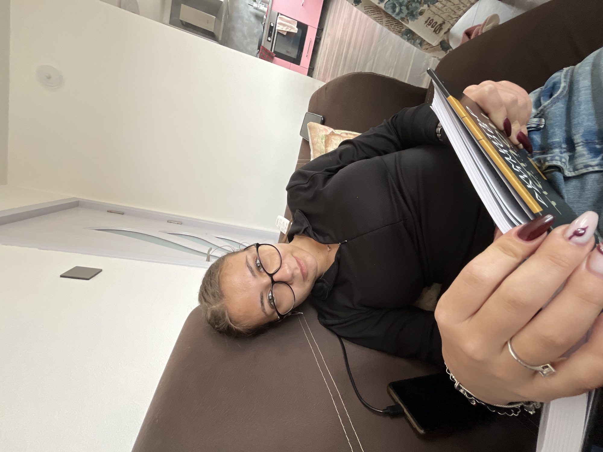 A young woman lies on a brown couch, holding a notebook. She wears a black hooded top and glasses. Her nails are long, with a dark red polish. A smartphone is beside her on the couch. The background includes a glimpse of a pink cabinet and an interior wall. The location is a room within an unspecified building in Karavelovo, Bulgaria.