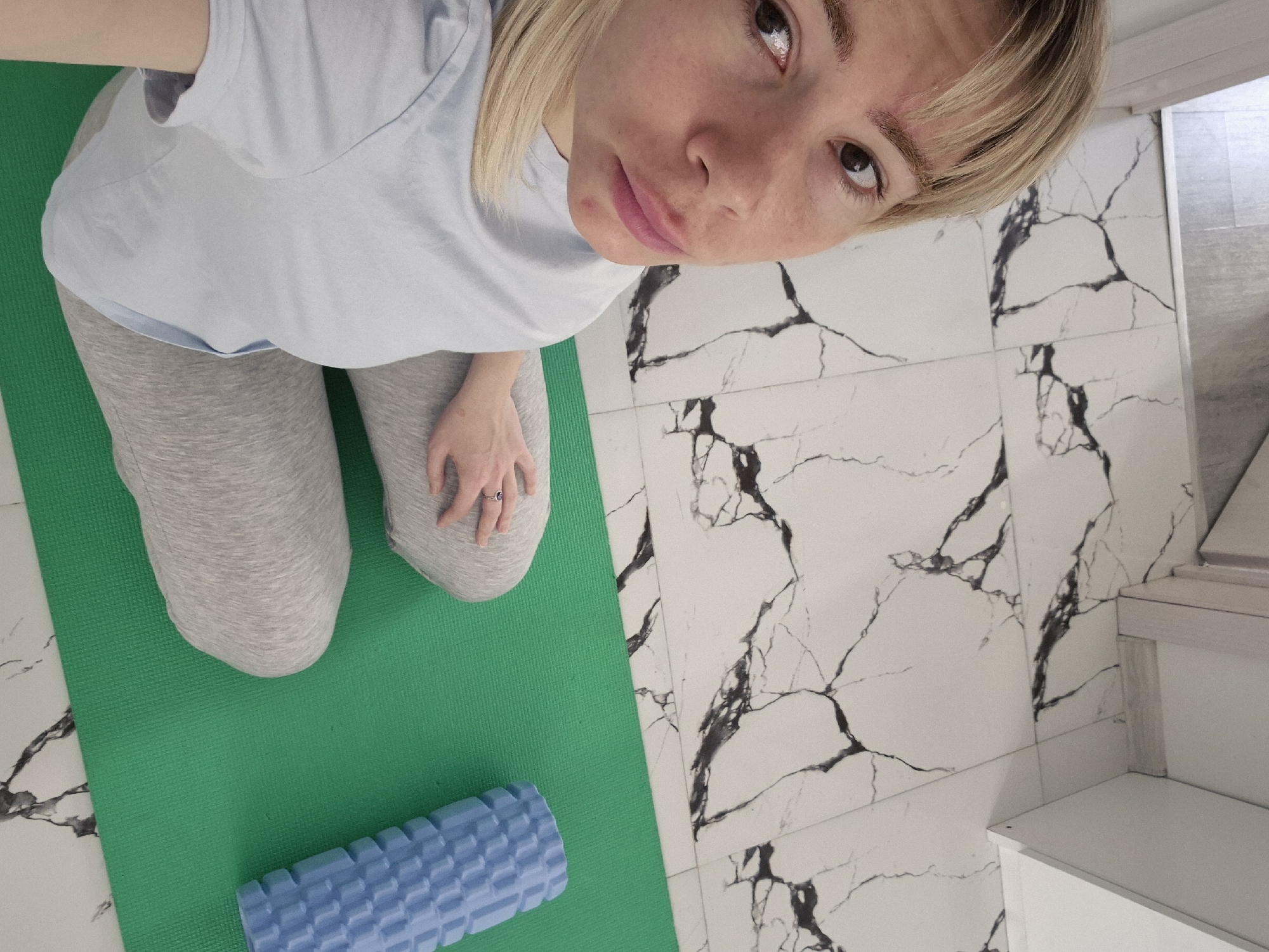 A woman kneels on a green yoga mat with a blue foam roller nearby, positioned on white tile flooring. The perspective is from above, looking down at the subject. The woman is wearing light gray sweatpants and a light blue t-shirt. The background shows marble-patterned white tiles and a portion of a white cabinet or shelf. The location is a room with tiled floors, possibly a bathroom or exercise area, inside a building in Batumi, Georgia. The exact location and address within Batumi cannot be determined from the image.