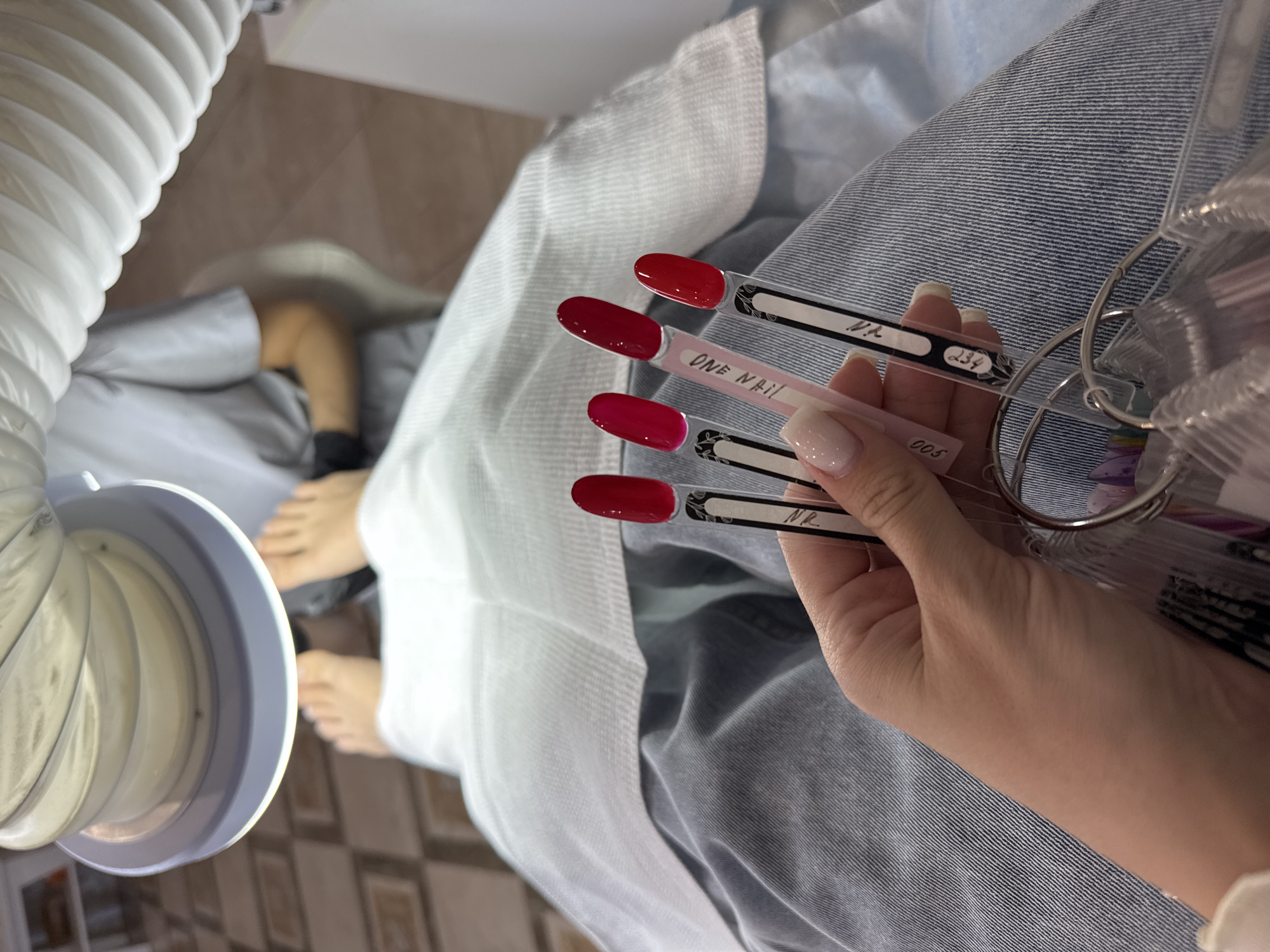 In a beauty salon in Minusinsk, Russia, a person's hand holds a ring of nail polish swatches. The swatches display various colors, with a focus on reds and a light nude shade. In the background, a person's feet are visible, positioned under a bright lamp, suggesting a pedicure service is in progress. The person receiving the service is wearing denim jeans and is draped with a white cloth. The environment appears to be a typical nail salon, with tiled flooring and professional equipment.