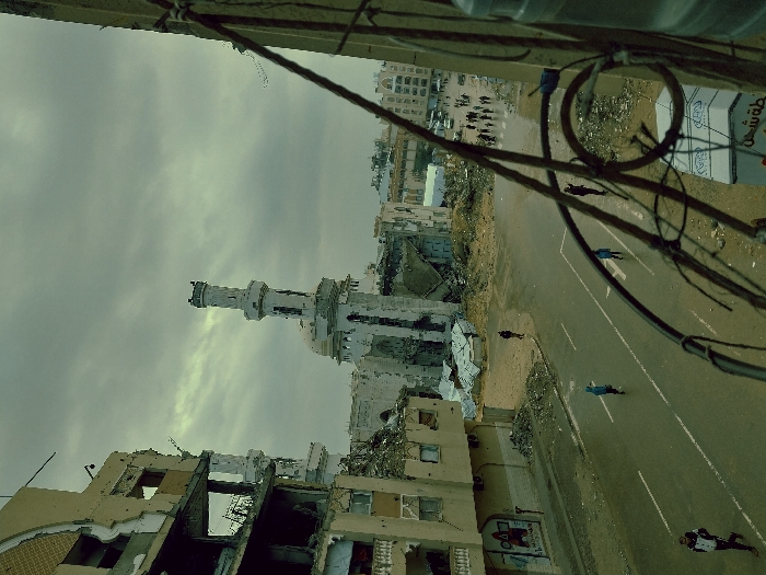 The image displays an elevated view of a street intersection in Khan Yunis, Palestinian Territory, characterized by extensive urban destruction under an overcast sky. In the background-center, a mosque with a tall, light-colored minaret stands, exhibiting visible damage to its dome and structure. To its left, a multi-story building is severely compromised, with large sections of its facade collapsed, exposing interior rooms and twisted rebar. Adjacent structures and the surrounding ground are covered in significant amounts of rubble and debris.
In the mid-ground, a paved road with faded white lane markings runs horizontally across the frame. Five individuals are walking on this road: one on the far left, two in blue garments closer to the center, and two others further right. On a cross-street in the distant right, a group of approximately fifteen pedestrians is visible, alongside several vehicles. The areas immediately bordering the road are covered in accumulated debris and loose earth. Graffiti is visible on a partially damaged building wall on the left side of the street.
The immediate foreground on the right side is partially obscured by dark, bent metal rods and wires, indicating the elevated perspective from a damaged structure. A portion of a white container or barrier with Arabic text is also visible in the lower right. The sky is uniformly gray and heavily clouded, contributing to the subdued lighting of the devastated urban landscape.