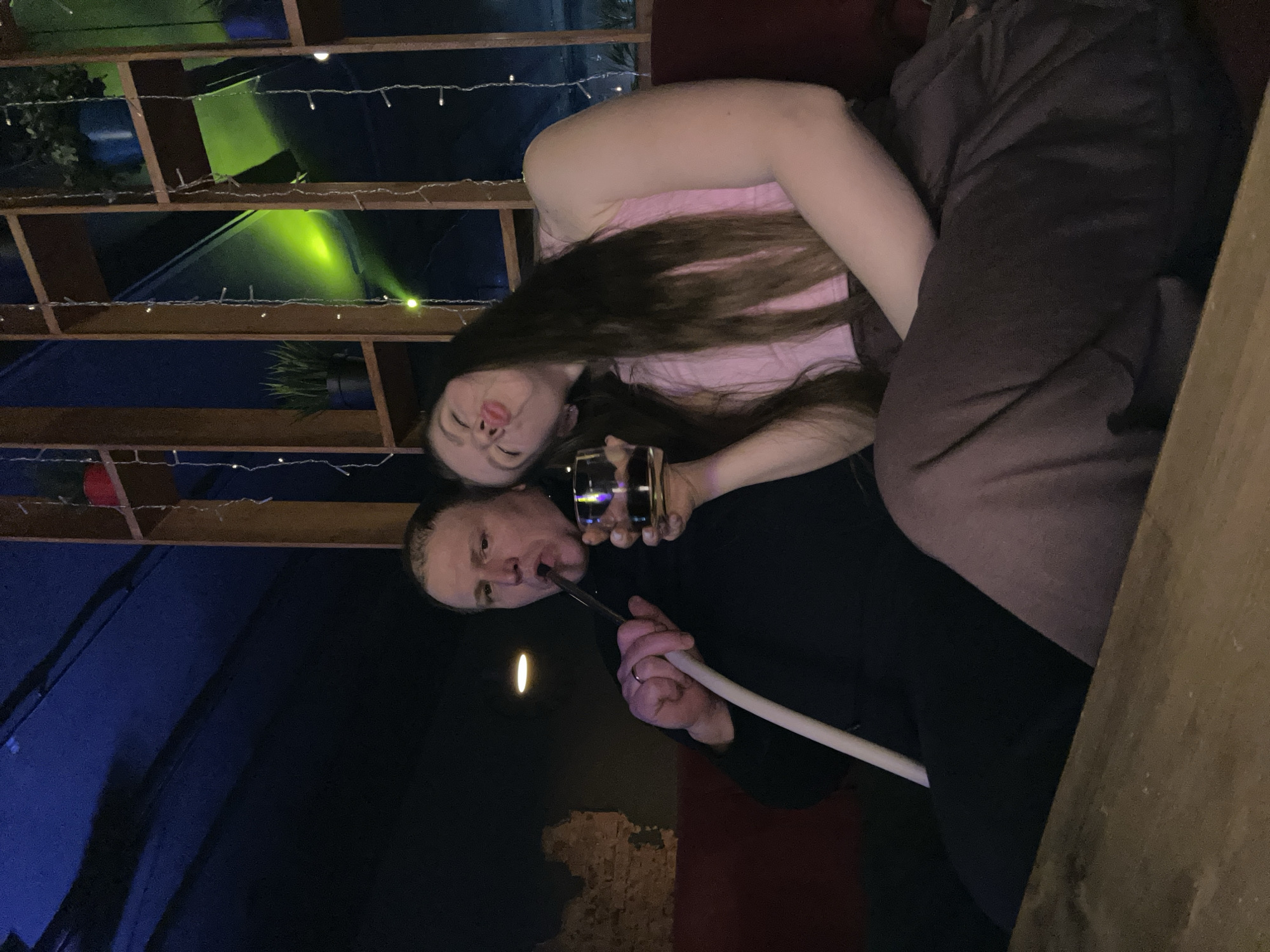 A man and woman are indoors at an establishment in Klin, Russia. The man is inhaling from a hookah hose. The woman is holding a glass. The interior is dimly lit with some decorative lights. A dark blue wall or backdrop is visible, as is a shelving unit with greenery and lights. The seating appears to be booth-style.
