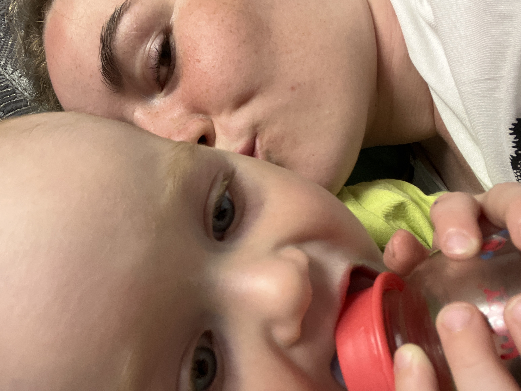 A close-up image shows a woman and a baby. The woman is lying down with her face close to the camera, her eyes closed. She has fair skin, dark eyebrows, and is wearing a white shirt. The baby is in the foreground, drinking from a baby bottle with a red nipple. The baby has fair skin and light-colored hair. The location is not discernible from the image. The scene involves the interaction between the woman and the baby, with the baby feeding.