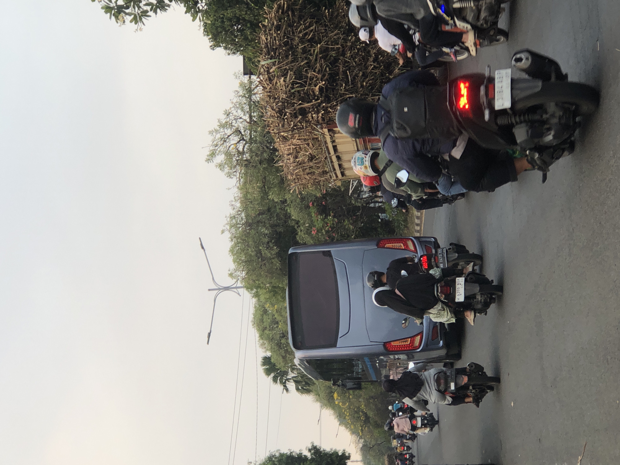 The image depicts a street scene in Ngaban, Indonesia, likely during the day due to the overcast sky. The foreground shows a busy road with multiple motorcycles and scooters. On the left, a grey scooter with a rider wearing a black hijab is visible. To its right, a blue bus is the dominant vehicle in the midground, its rear facing the camera. Behind the bus, another scooter carries two individuals, one wearing a black hijab and the other a black shirt. Further down the road, more motorcycles and riders are present, some carrying passengers. The right side of the image features a large truck overloaded with dry, woody materials, possibly sugar cane stalks, suggesting transport for agricultural or industrial purposes. The surrounding environment includes trees and foliage lining the street, with streetlights visible in the distance. The overall impression is one of urban traffic congestion in a tropical or subtropical setting.