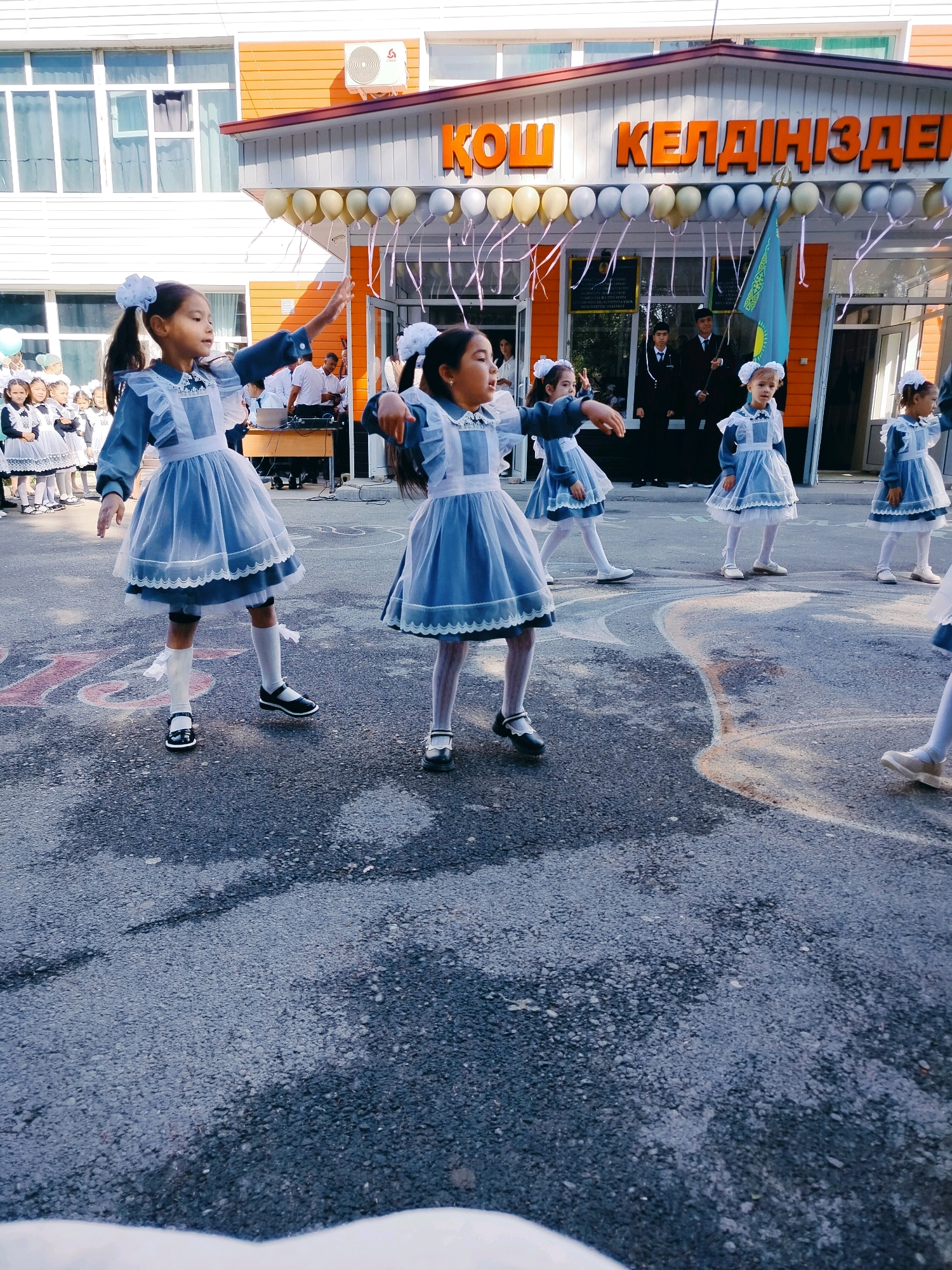 The image captures a formal outdoor event in front of a school building. Several young girls, dressed in matching blue uniforms with white collars and trim, are performing a synchronized dance on an asphalt surface. The girls have their hair styled with white bows. In the background, the school building is visible with an orange facade and a large sign bearing text in Cyrillic script, which is identified as being from the city of Kölkent, Kazakhstan. Balloons in shades of yellow, gold, and white are strung across the entrance. A flag of Kazakhstan is also present, held by an individual in the background, along with other adults and children observing the event. The lighting suggests it is daytime, likely morning or early afternoon.